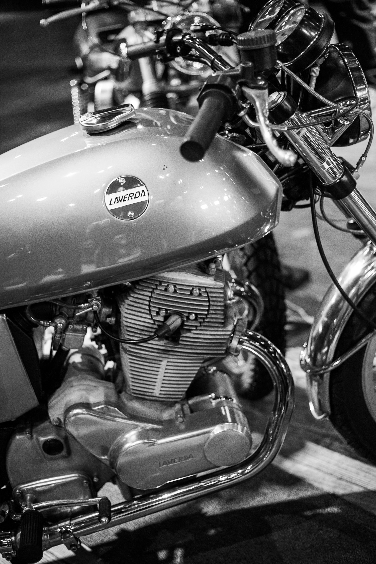 A high-contrast vertical black and white photograph of a vintage Laverda motorcycle, detailing the engine block, chrome exhaust pipe, and fuel tank with emblem