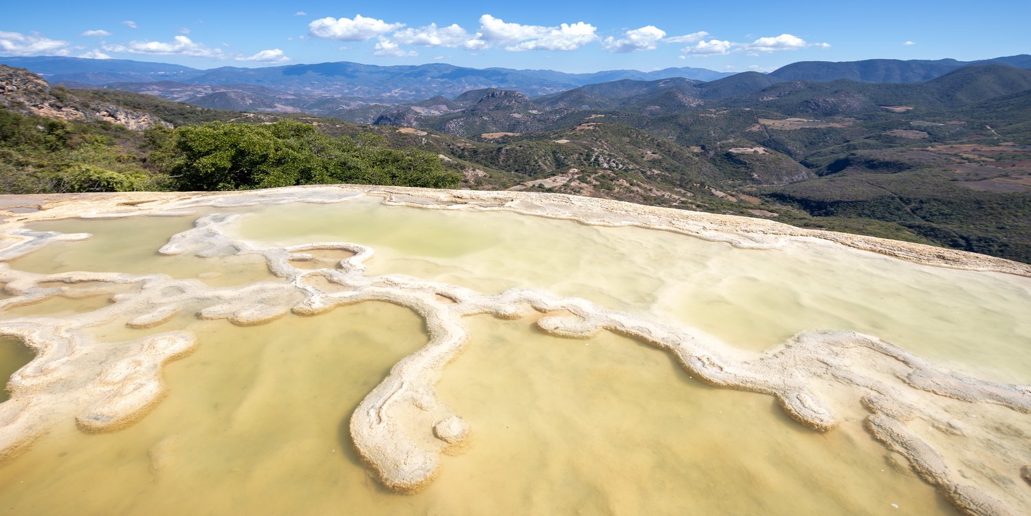 A wide-angle landscape photograph looking across the pale yellow and white calcium mineral pools of Hierve el Agua, showcasing the rugged green mountains and blue sky of Oaxaca, Mexico.