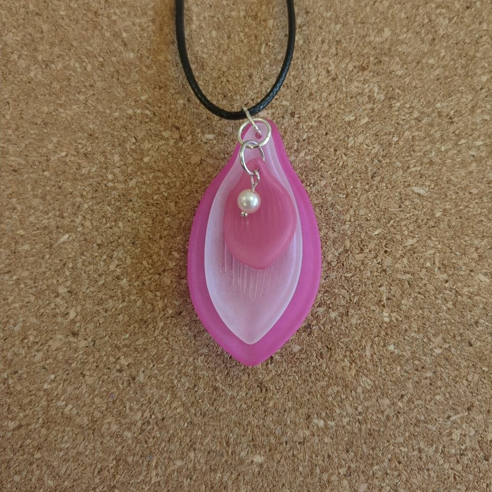 A pendant made of three stacked petal shapes in different shades of pink. a pearl detail sits at the top of the innermost petal. The petals are connected with silver toned metal rings and strung on a black cord.
