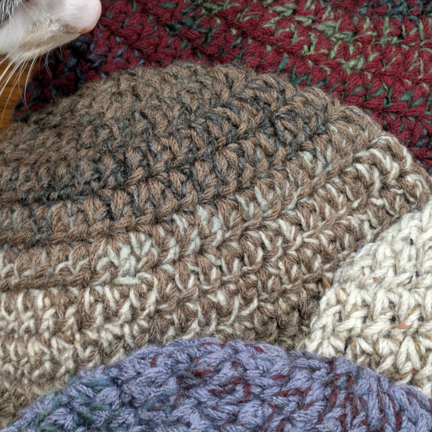 Close-up of hands crocheting a chunky double crochet beanie with variegated brown, gray, black, and cream yarn using a green crochet hook, with a calico cat blurred in the background.