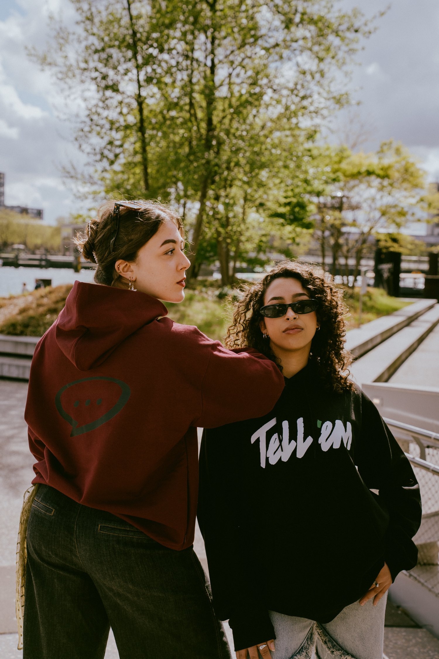 Foto van twee vrouwen die samen buiten staan. De vrouw links staat met haar rug naar de camera gedraait,en leunt op de schouder van de andere vrouw. De rechterkant van haar gezicht en haar rug zijn duidelijk zichtbaar en ze draagt de Spraakuhloos Premium Hoodie in de kleur Wine met een groot zwart Spraakuhloos logo op de rug. De vrouw rechts heeft een zwarte zonnebril op en kijkt direct in de camera. Zij draagt de Tell 'em Basic Hoodie in de kleur Black met een groot wit Tell 'em logo op de rug. Op de achtergrond zie je bomen, wolken, water en gebouwen.