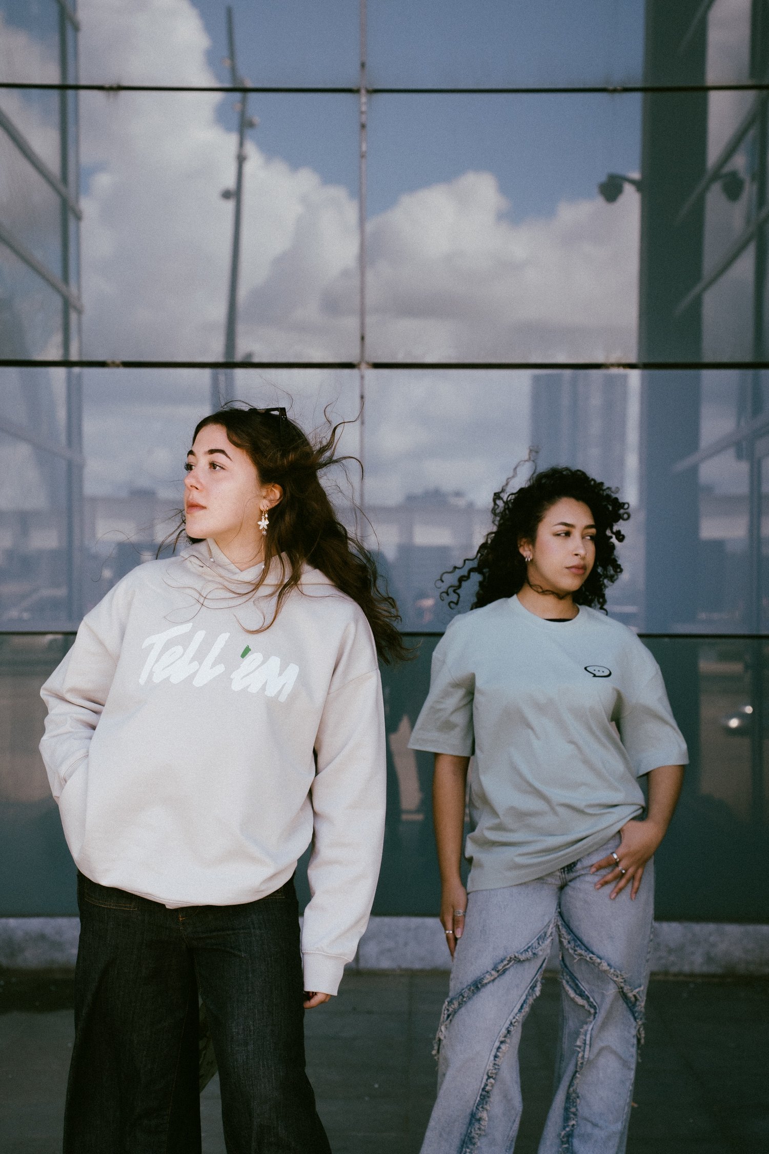 Foto van twee vrouwen die voor de glazen wand van een gebouw staan en beiden een andere richting in de verte kijken. De vrouw links heeft lang bruin haar en draagt een Tell 'em Premium Hoodie in de kleur Clay met een groot wit Tell 'em logo op de borst. De vrouw rechts draagt een Spraakuhloos T-shirt in de kleur Sage met een klein zwart Spraakuhloos logo op de borst. Op de achtergrond zie je dat de glazen wand van het gebouw wolken en een deel van een skyline reflecteert.