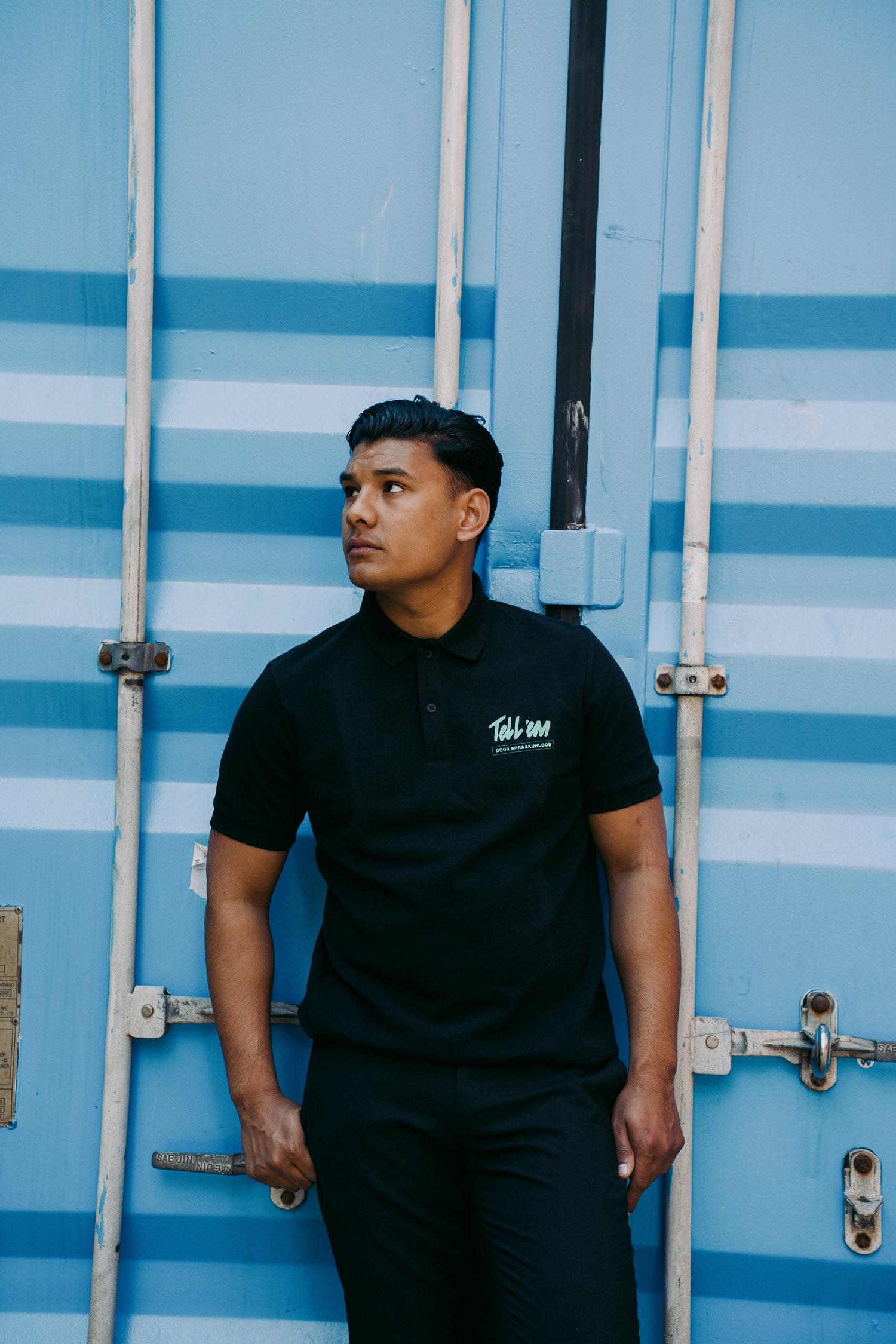 Foto van een man met kort zwart haar voor een blauwe container die in de verte kijkt. Hij draagt het zwarte Tell 'em Poloshirt met een klein turquoise Tell 'em logo op de borst.