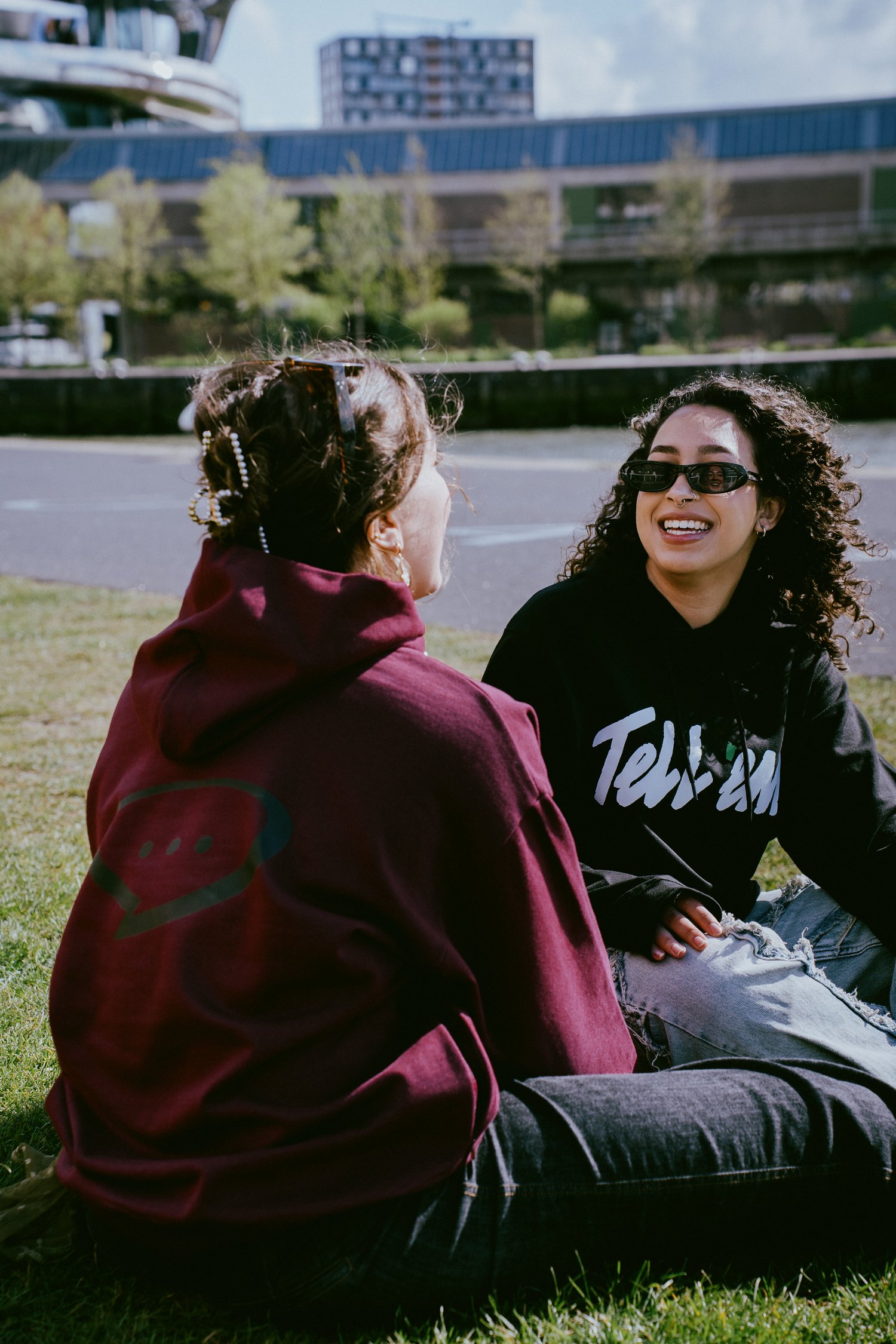 Foto van twee vrouwen die schuin tegenover elkaar op een grasveld voor het water zitten. De vrouw links zit met haar rug naar de camera gedraait en kijkt naar haar vriendin. Ze draagt de Spraakuhloos Premium Hoodie in de kleur Wine met een groot zwart Spraakuhloos logo op de rug dat nog net zichtbaar is. De vrouw rechts heeft een zwarte zonnebril op en is zichtbaar lachend aan het praten. Zij draagt de Tell 'em Basic Hoodie in de kleur Black met een groot wit Tell 'em logo op de borst. Op de achtergrond zie je bomen, wolken, water en gebouwen.