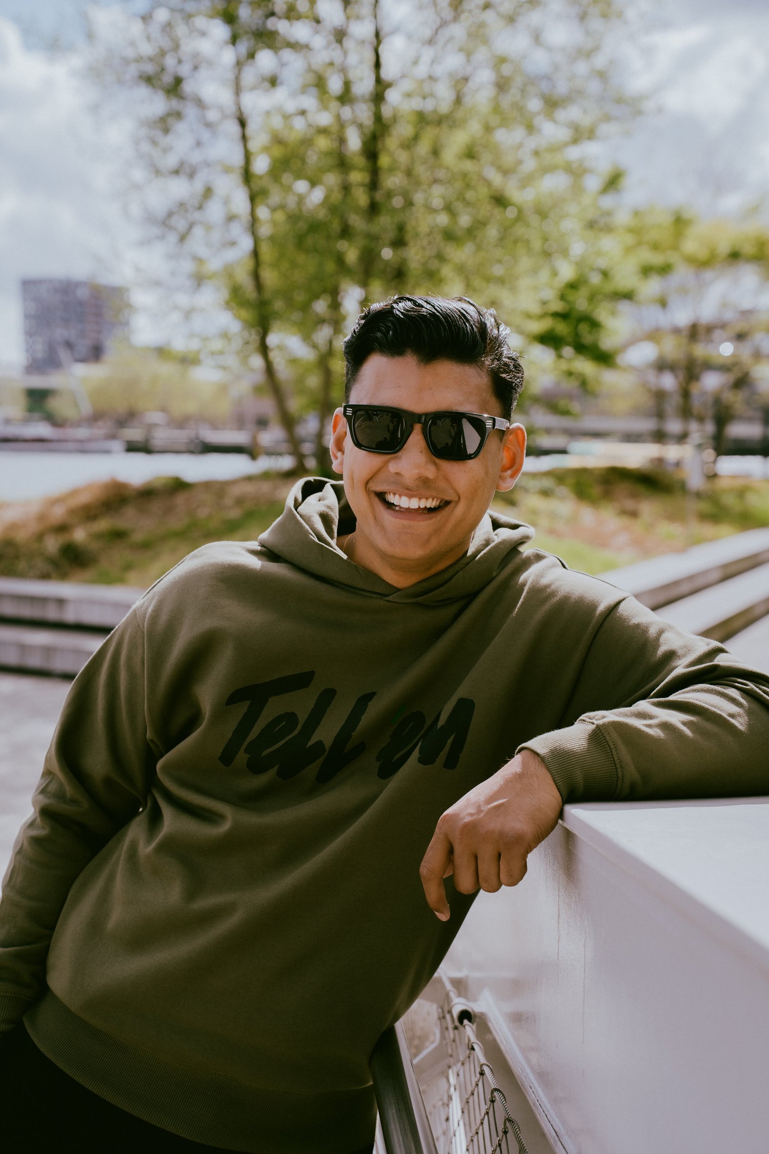 Foto van een man met kort zwart haar en een zwarte zonnebril, die met een arm leunt op een brede witte railing en lachend in de camera kijkt. Hij draagt de Tell 'em Premium Hoodie in de kleur Light Khaki met een groot zwart Tell 'em logo op de borst. Op de achtergrond zie je bomen, wolken, water en gebouwen.