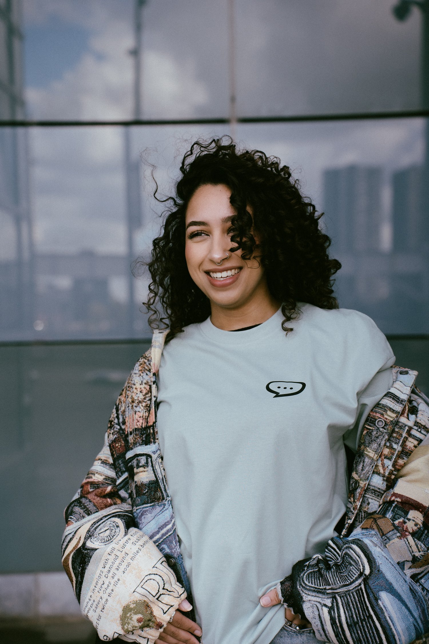Foto van een lachende vrouw met krullend zwart haar die voor de glazen wand van een gebouw staat en in de verte kijkt. Ze draagt een Spraakuhloos T-shirt in de kleur Sage met een klein zwart Spraakuhloos logo op de borst. Over het shirt draagt ze een lichtgekleurde bomberjack met een grafische print. Op de achtergrond zie je dat de glazen wand van het gebouw wolken en een deel van een skyline reflecteert.
