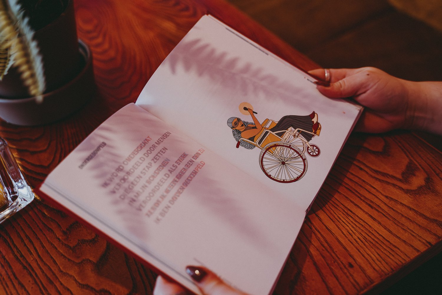Open boek op een houten tafel. Op de linkerpagina staat een anaglyf gedicht in het Nederlands. Op de rechterpagina staat een illustratie van een persoon in een rolstoel, ze houdt een microfoon vast in haar hand.