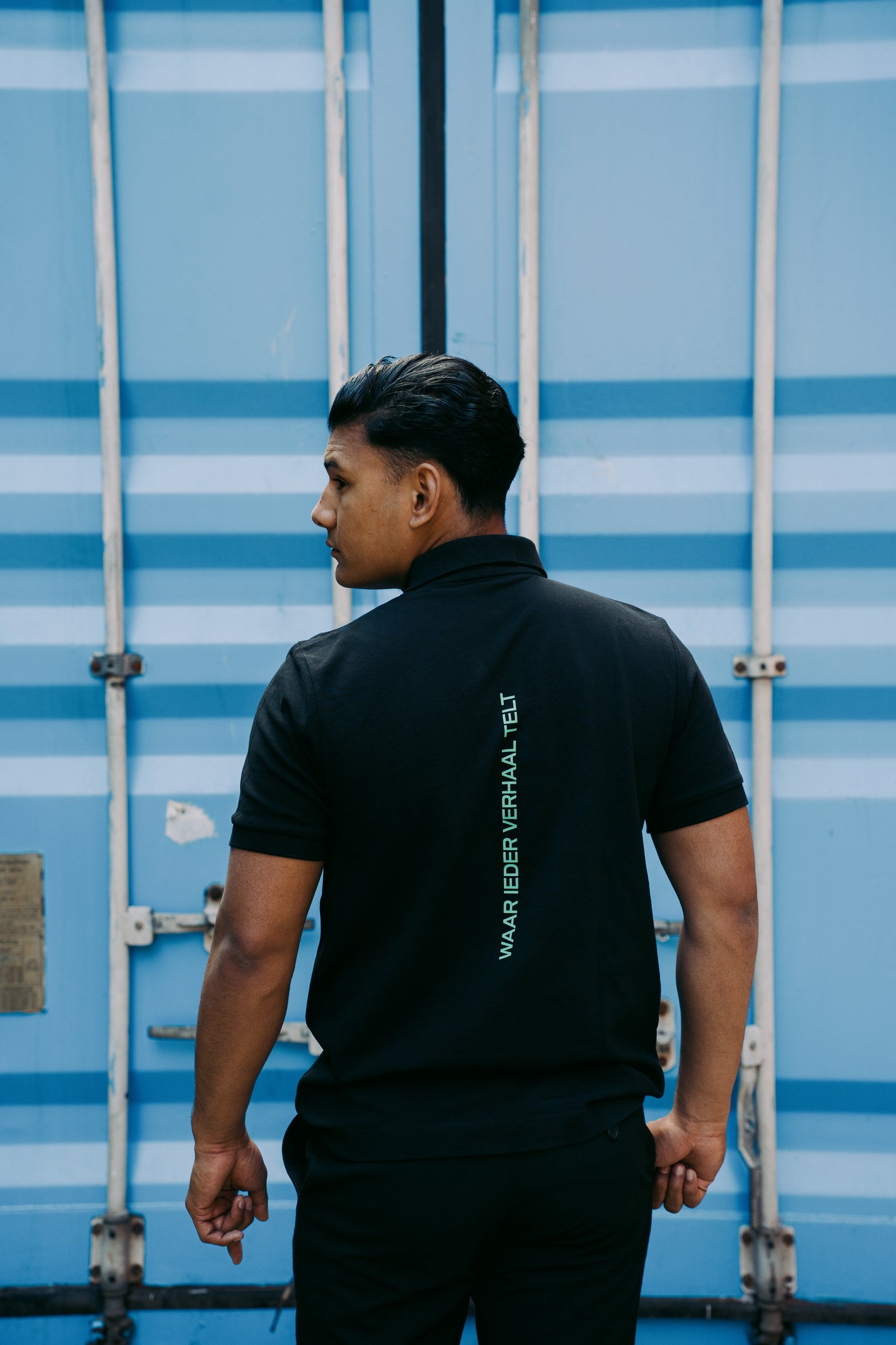 Foto van een man met kort zwart haar voor een blauwe container met zijn rug schuin naar de camera. Hij draagt het zwarte Tell em Poloshirt met de turquoise gekleurde boodschap "Waar ieder verhaal telt" verticaal over de ruggengraat.