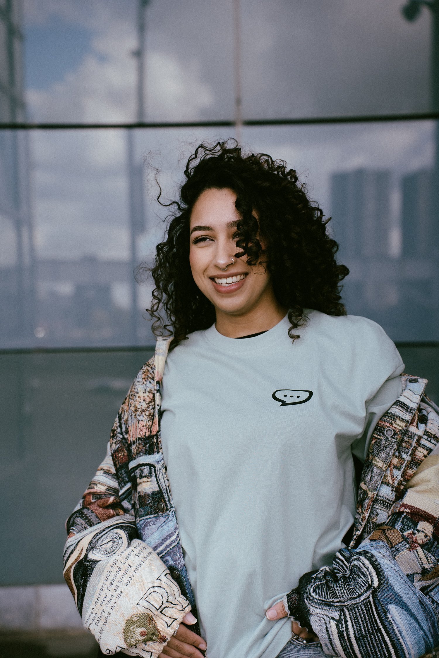 Foto van een lachende vrouw met krullend zwart haar die voor de glazen wand van een gebouw staat en in de verte kijkt. Ze draagt een Spraakuhloos T-shirt in de kleur Sage met een klein zwart Spraakuhloos logo op de borst. Over het shirt draagt ze een lich