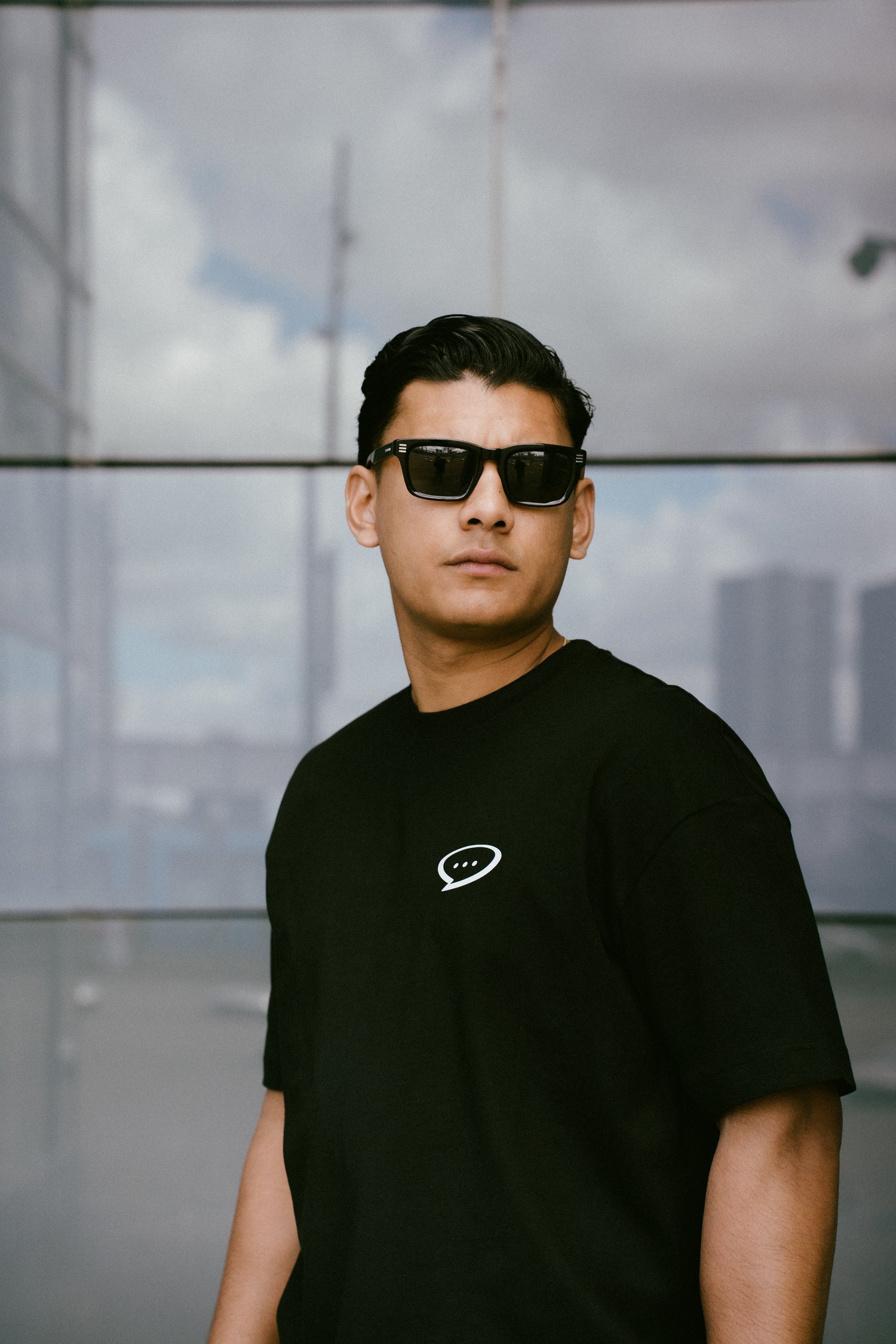 Foto van een man met kort zwart haar en een zwarte zonnebril die voor de glazen wand van een gebouw staat en in de verte kijkt. Hij draagt een Spraakuhloos T-shirt in de kleur Black met een klein wit Spraakuhloos logo op de borst. Op de achtergrond zie je