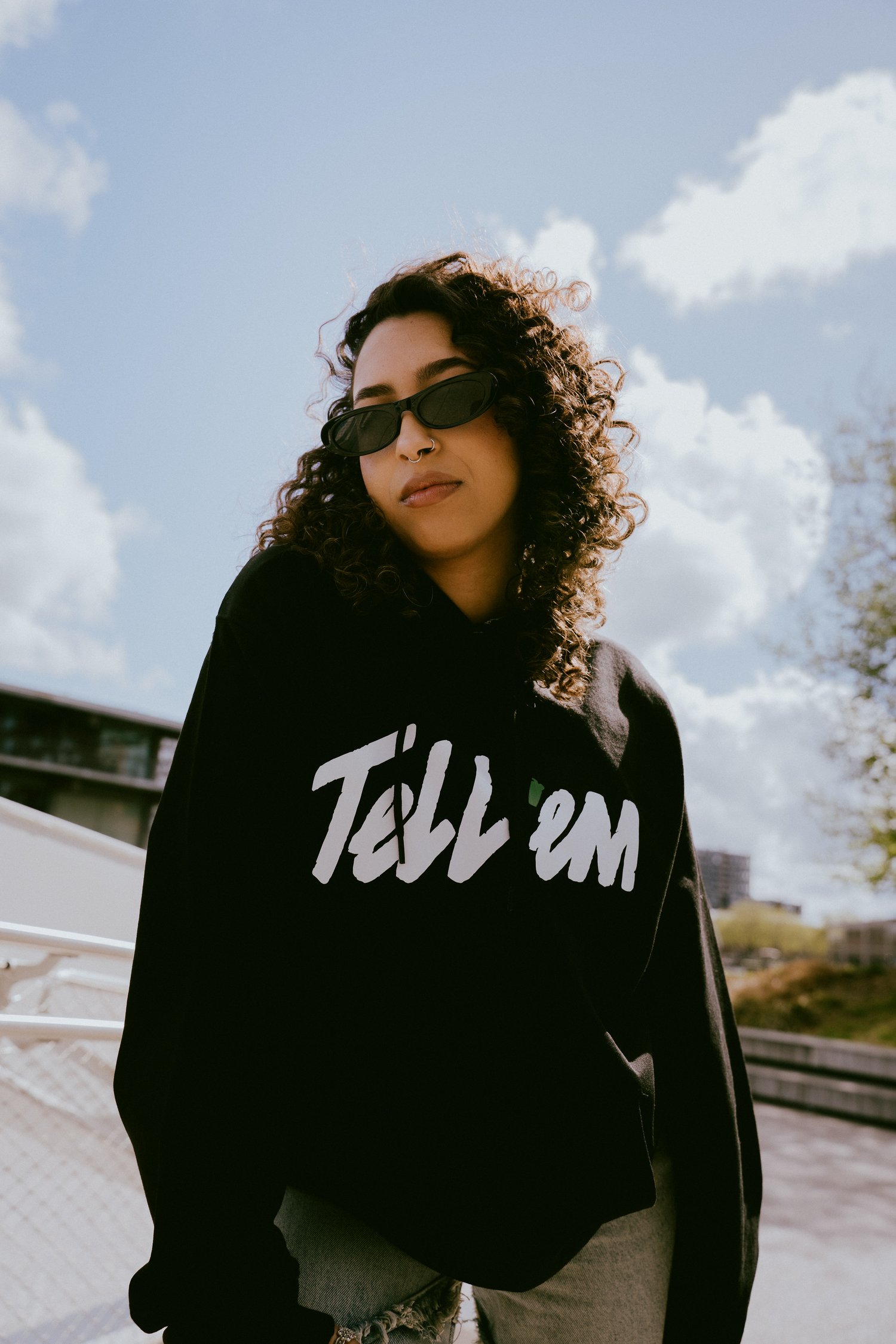 Foto van een vrouw met krullend zwart haar en een zwarte zonnebril. Ze staat enigszins voorover gebogen en kijkt direct en zelfverzekerd in de camera. Ze draagt de Tell 'em Basic Hoodie in de kleur Black met een groot wit Tell 'em logo op de borst. Op de 