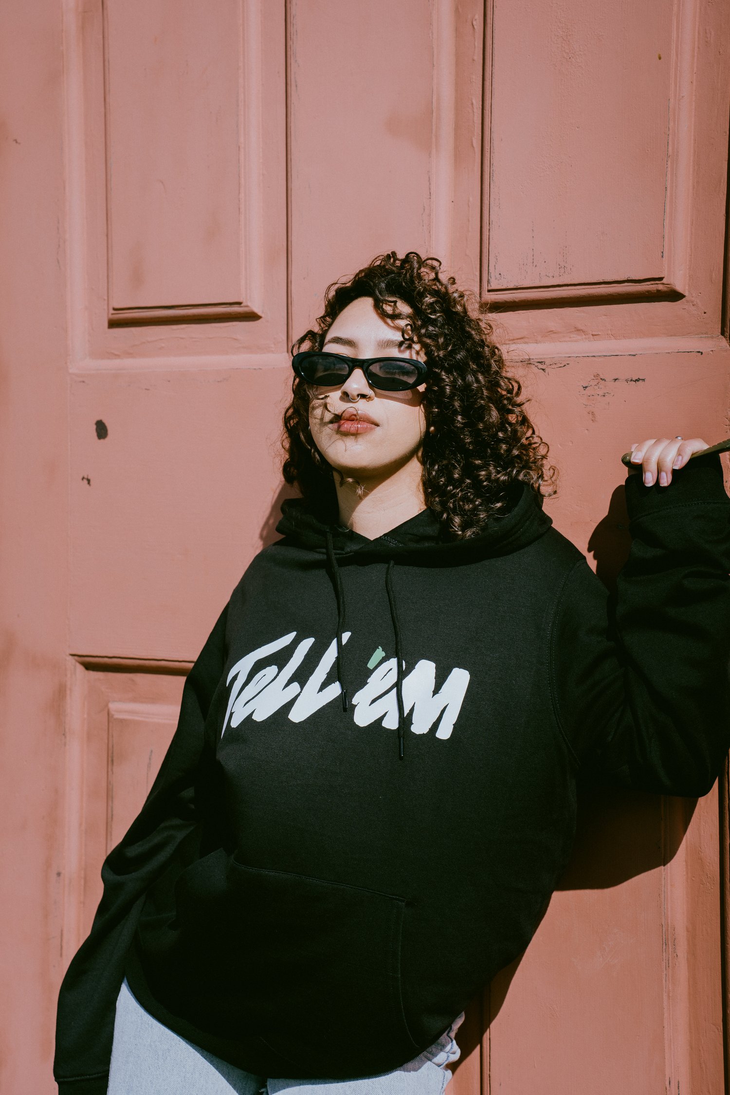 Foto van een vrouw met zwart krullend haar en een zwarte zonnebril, die voor een terracotta gekleurde deur staat. Ze kijkt direct in de camera en draagt de Tell 'em Basic Hoodie in kleur Black met een groot wit Tell 'em logo op de borst.