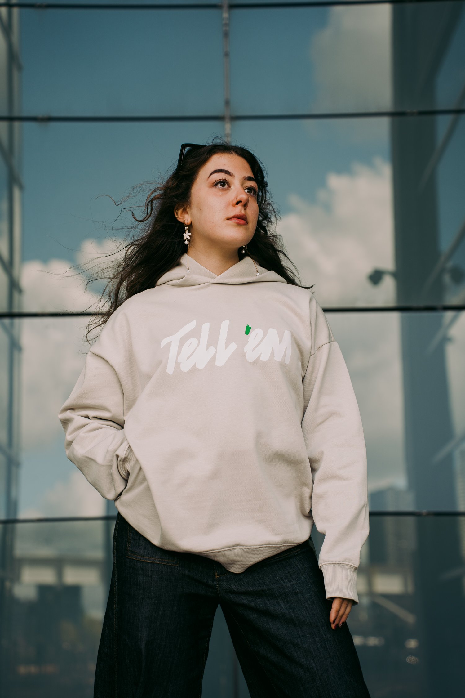 Foto van een vrouw met lang bruin haar die voor de glazen wand van een gebouw staat en in de verte kijkt. Ze draagt een Tell em Premium Hoodie in de kleur Clay met een groot wit Tell em logo op de borst. Ze heeft haar rechterhand in de onzichtbare zijza