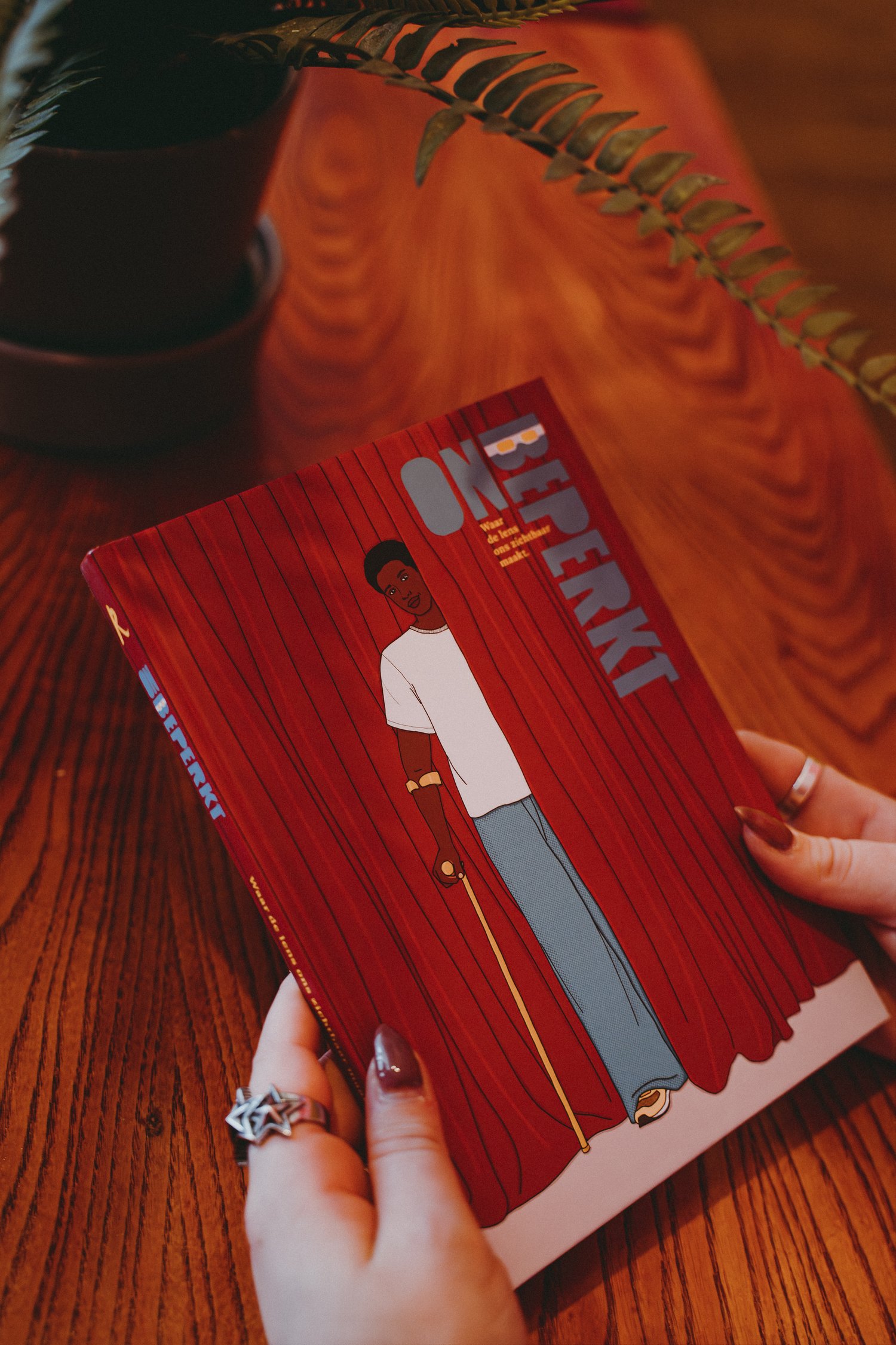 Boek met de titel “Onbeperkt” wordt door twee handen vast gehouden boven een houten tafel. De omslag van het boek toont een illustratie van een persoon voor rode gordijnen met een gele wandelstok.