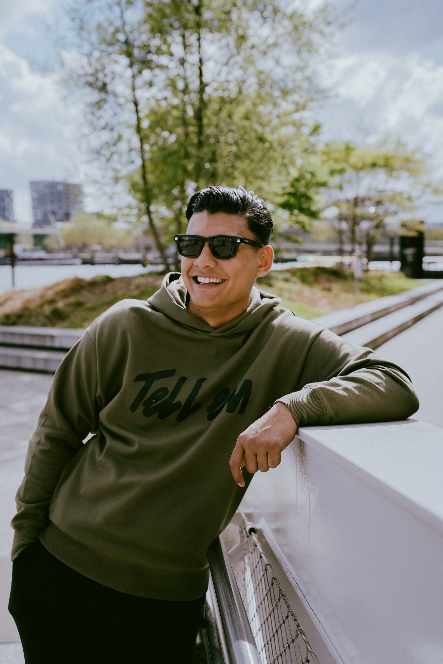 Foto van een man met kort zwart haar en een zwarte zonnebril, die met een arm leunt op een brede witte railing en lachend in de verte kijkt. Hij draagt de Tell em Premium Hoodie in de kleur Light Khaki met een groot zwart Tell em logo op de borst. Op de