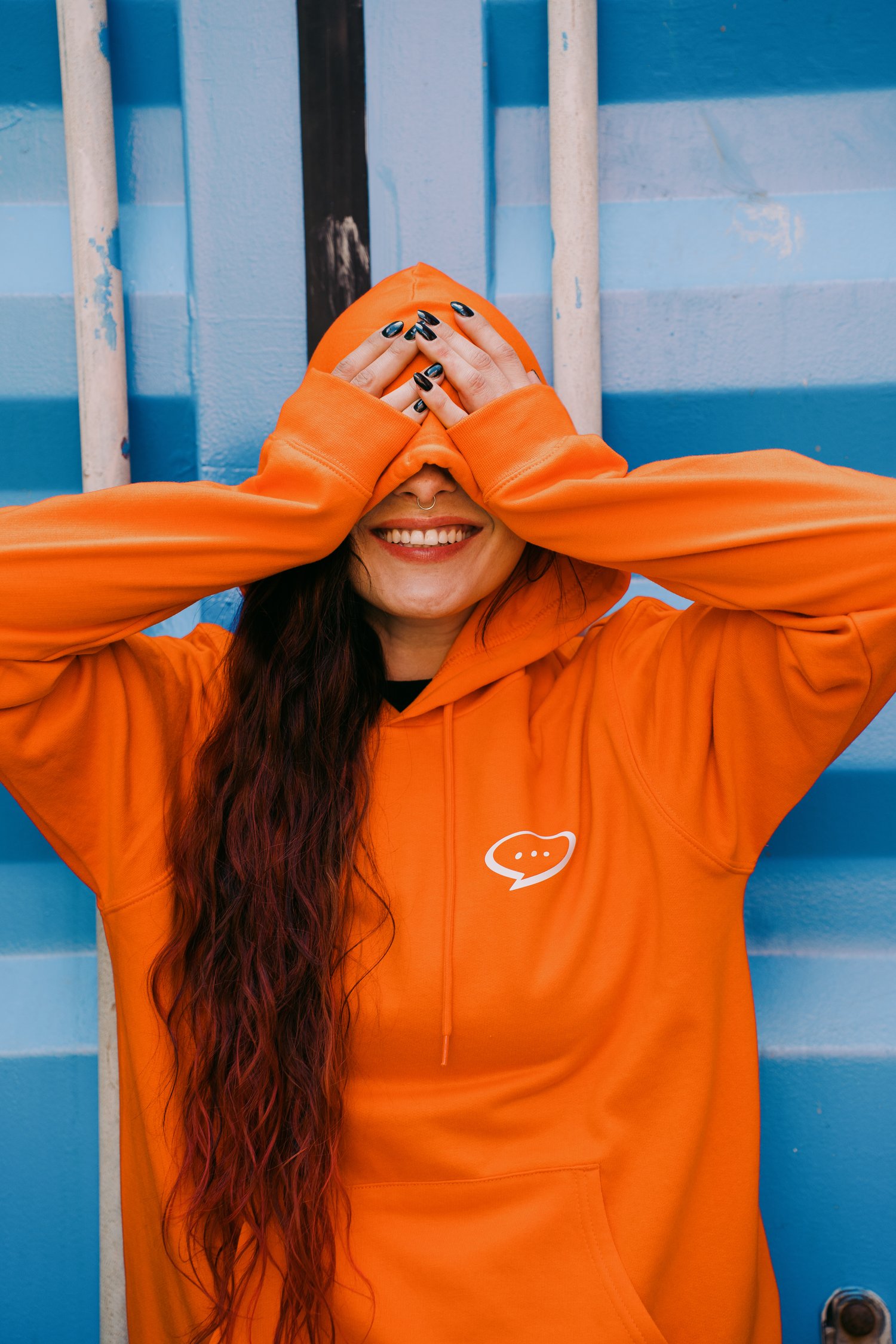 Foto van een lachende vrouw met lang roodbruin haar. Ze draagt de Spraakuhloos Basic Hoodie in de kleur Orange met een klein wit Spraakuhloos logo op de borst. Ze bedekt haar ogen met haar capuchon en beide handen en staat voor een blauwe achtergrond.