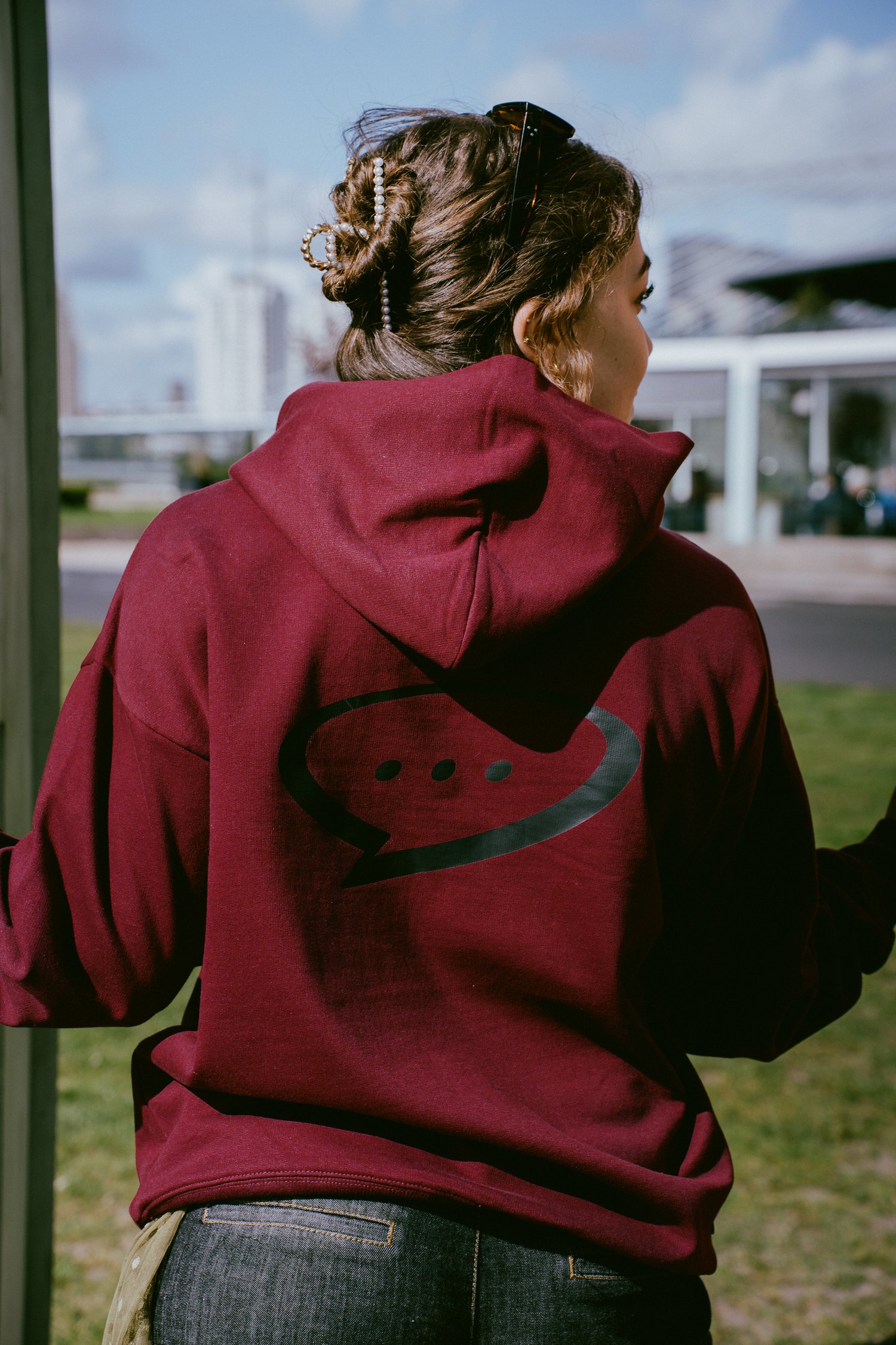 Foto van de achterkant van de Spraakuhloos Premium Hoodie in de kleur Wine. Het model heeft bruin opgestoken haar en staat met haar rug naar de camera. Op de achtergrond zie je een grasveld en een skyline.