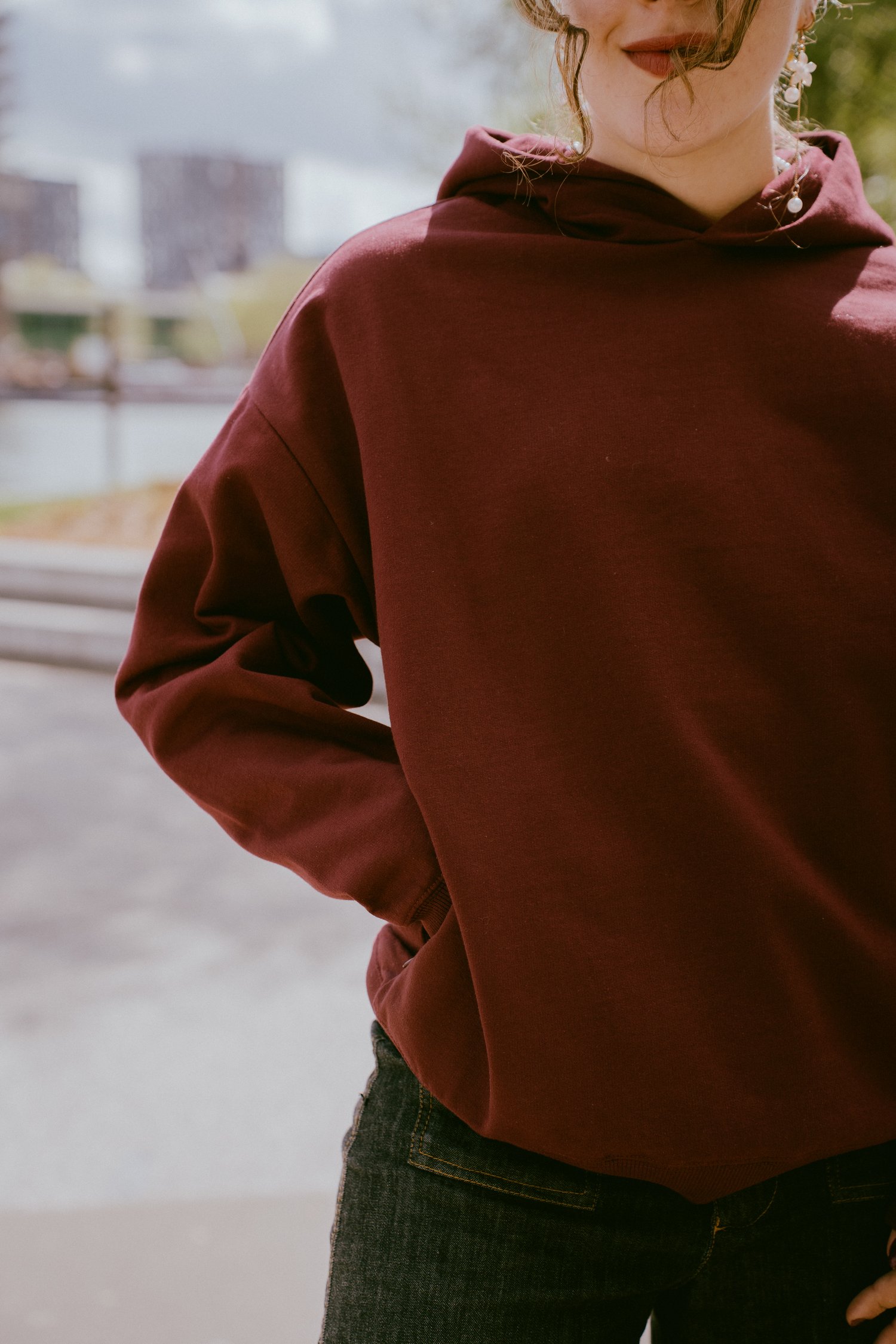 Close up foto van de voorkant van de Spraakuhloos Premium Hoodie in de kleur Wine. Het model heeft haar hand in de onzichtbare zijzak van de hoodie. Op de achtergrond zie je bomen, gebouwen, wolken en water.