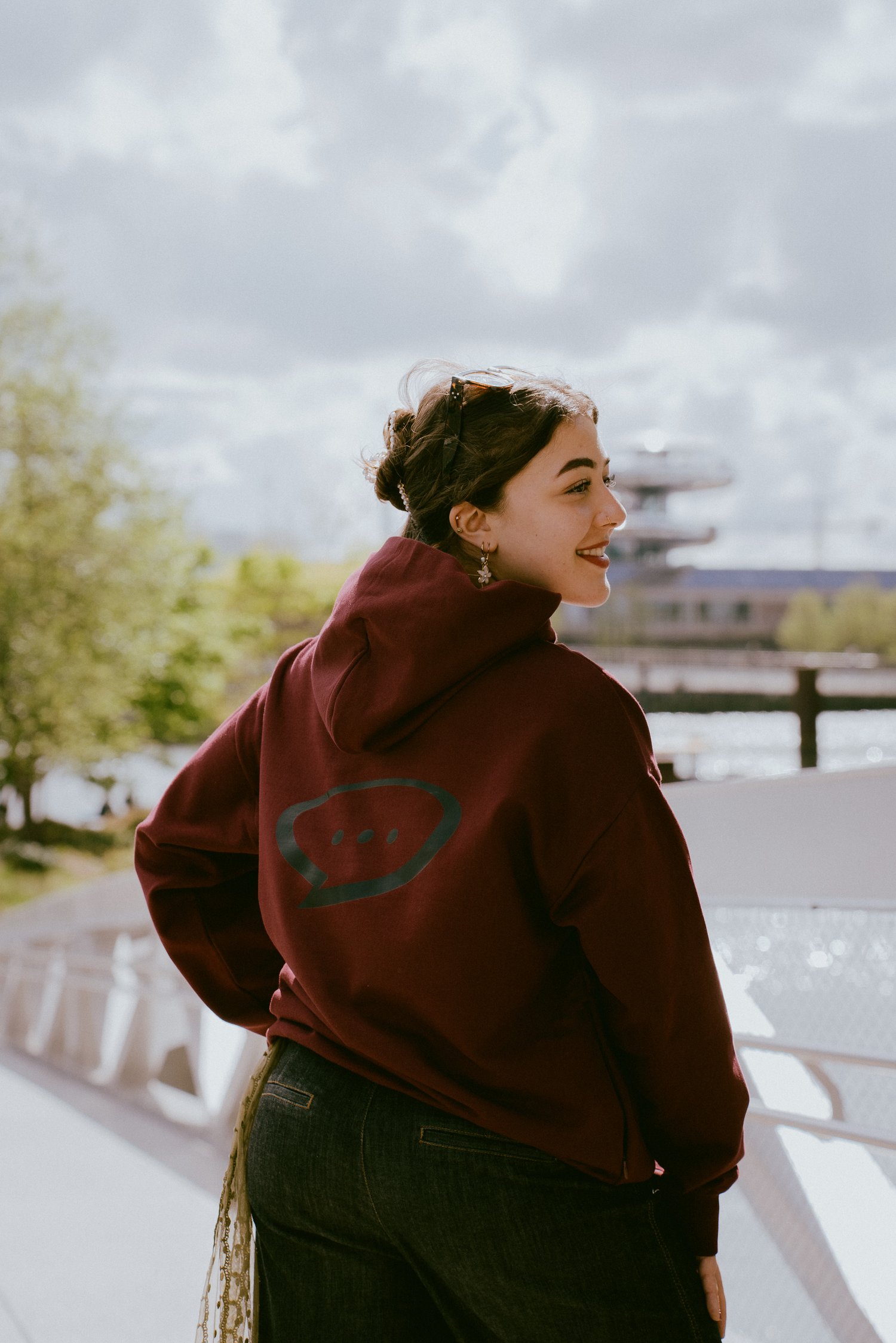 Foto van een glimlachende vrouw die met haar rug naar de camera gedraait staat. De rechterkant van haar gezicht en haar rug zijn duidelijk zichtbaar. Ze draagt een Spraakuhloos Premium Hoodie in de kleur Wine met een groot zwart Spraakuhloos logo op de ru