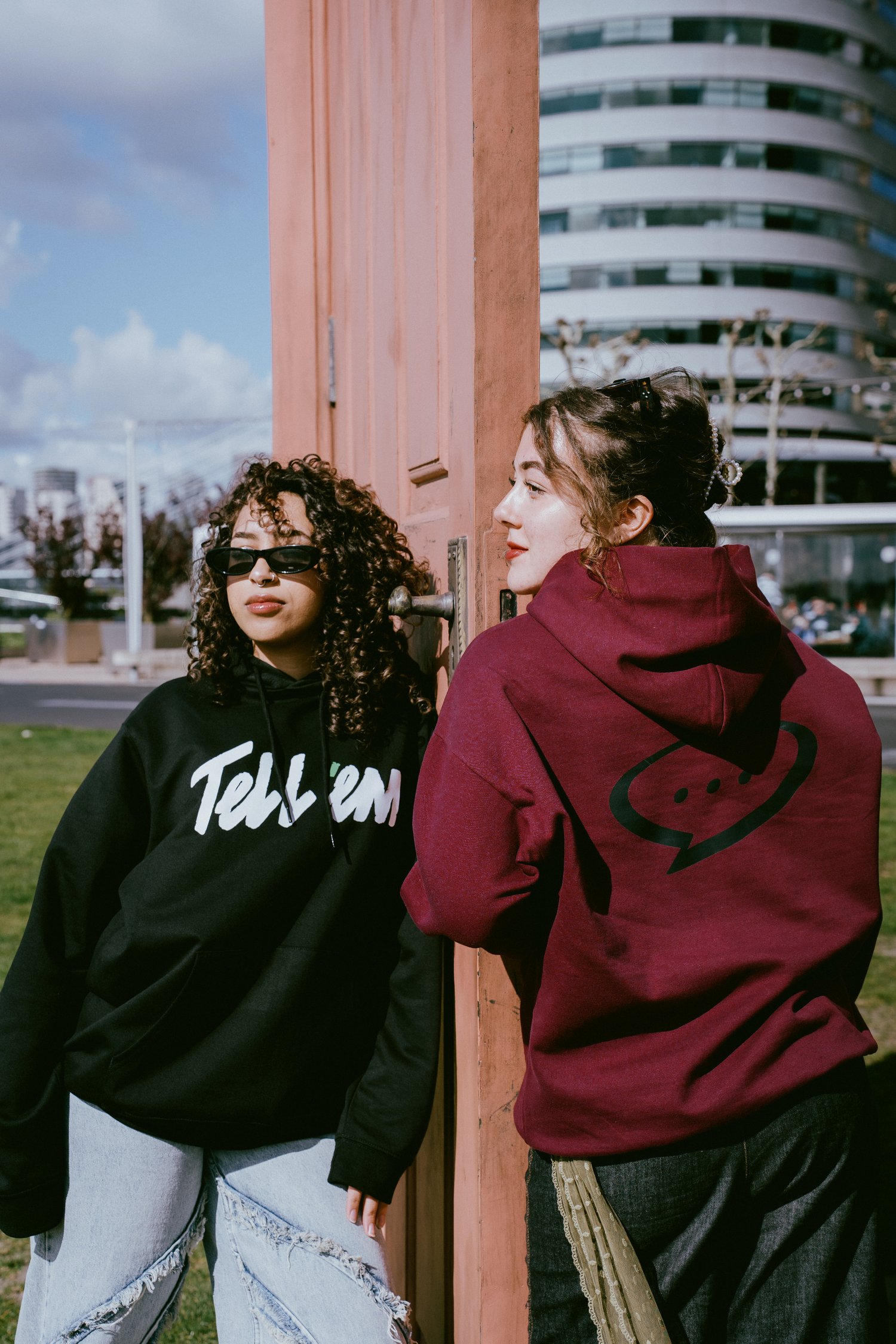 Foto van twee vrouwen die aan beide kanten van een zwevende terracotta gekleurde deur leunen. De vrouw links heeft een zwarte zonnebril op en kijkt in de verte. Zij draagt de Tell em Basic Hoodie in de kleur Black met een groot wit Tell em logo op de bo