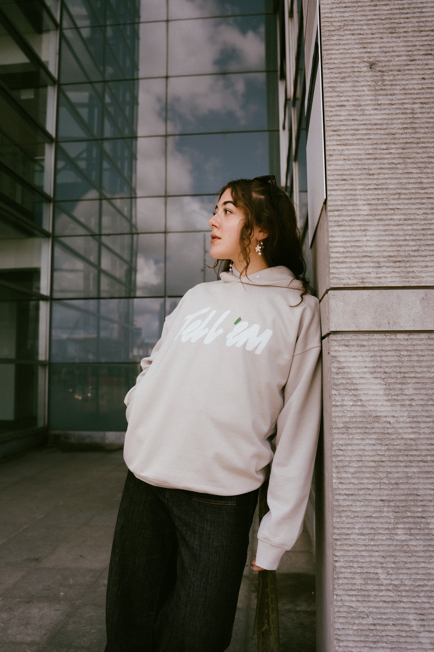 Foto van een vrouw met lang bruin haar die tegen een stenen muur van een gebouw aanleunt en in de verte kijkt. Ze draagt een Tell em Premium Hoodie in de kleur Clay met een groot wit Tell em logo op de borst. Op de achtergrond zie je dat de glazen wand
