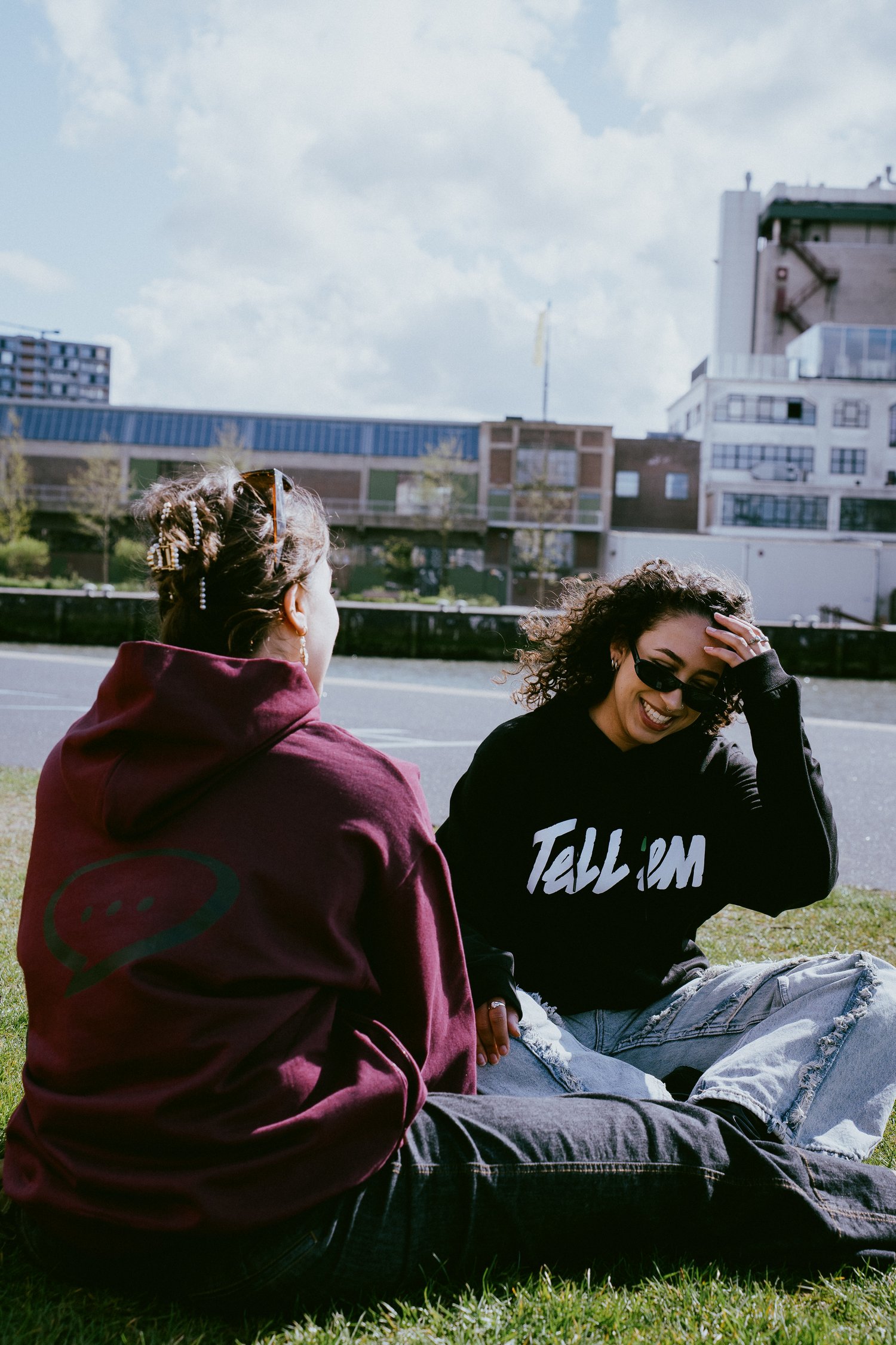 Foto van twee vrouwen die schuin tegenover elkaar op een grasveld voor het water zitten. De vrouw links zit met haar rug naar de camera gedraait en kijkt naar haar vriendin. Ze draagt de Spraakuhloos Premium Hoodie in de kleur Wine met een groot zwart Spr