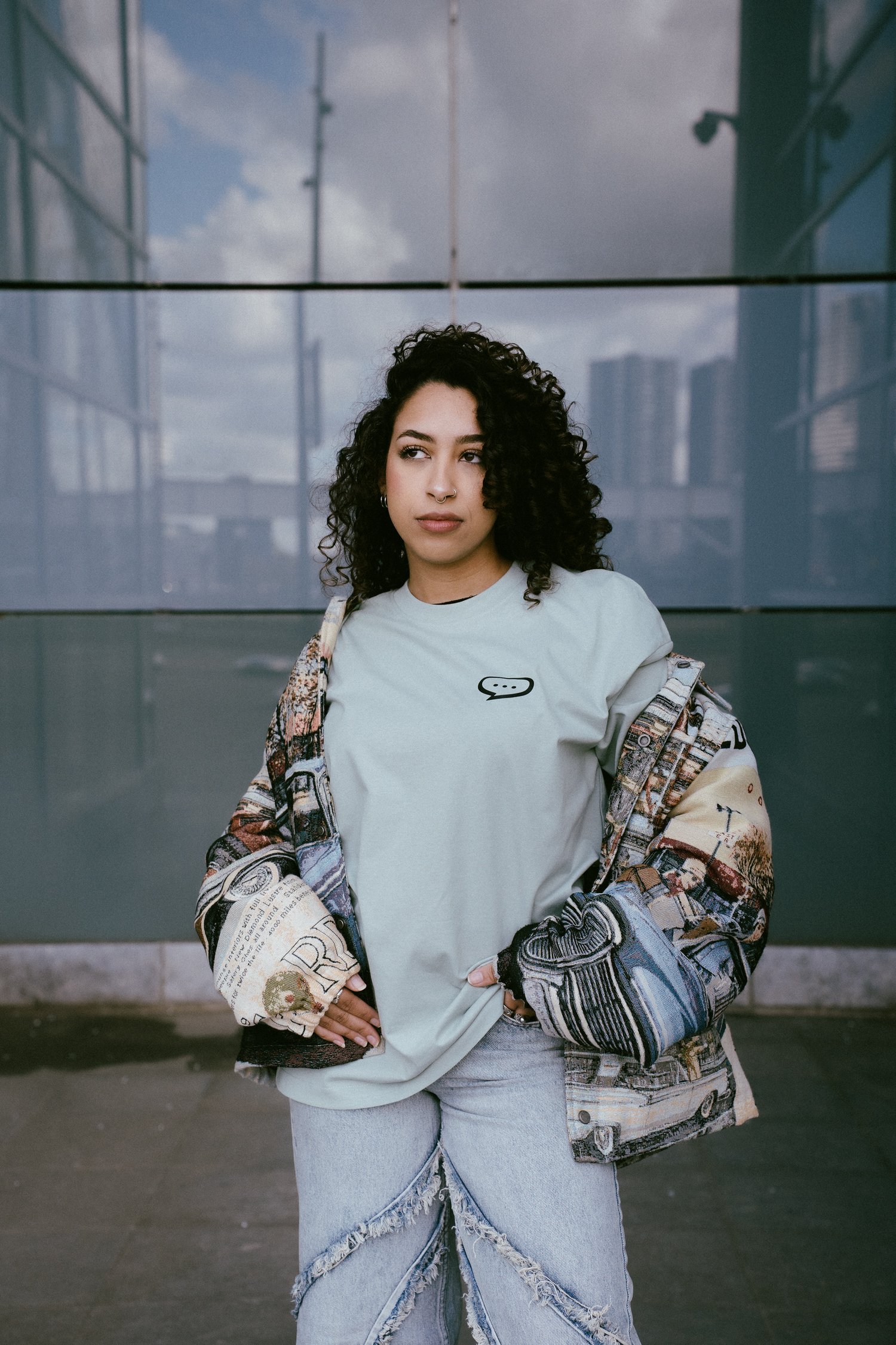 Foto van een vrouw met krullend zwart haar die voor de glazen wand van een gebouw staat en in de verte kijkt. Ze draagt een Spraakuhloos T-shirt in de kleur Sage met een klein zwart Spraakuhloos logo op de borst. Over het shirt draagt ze een lichtgekleurd