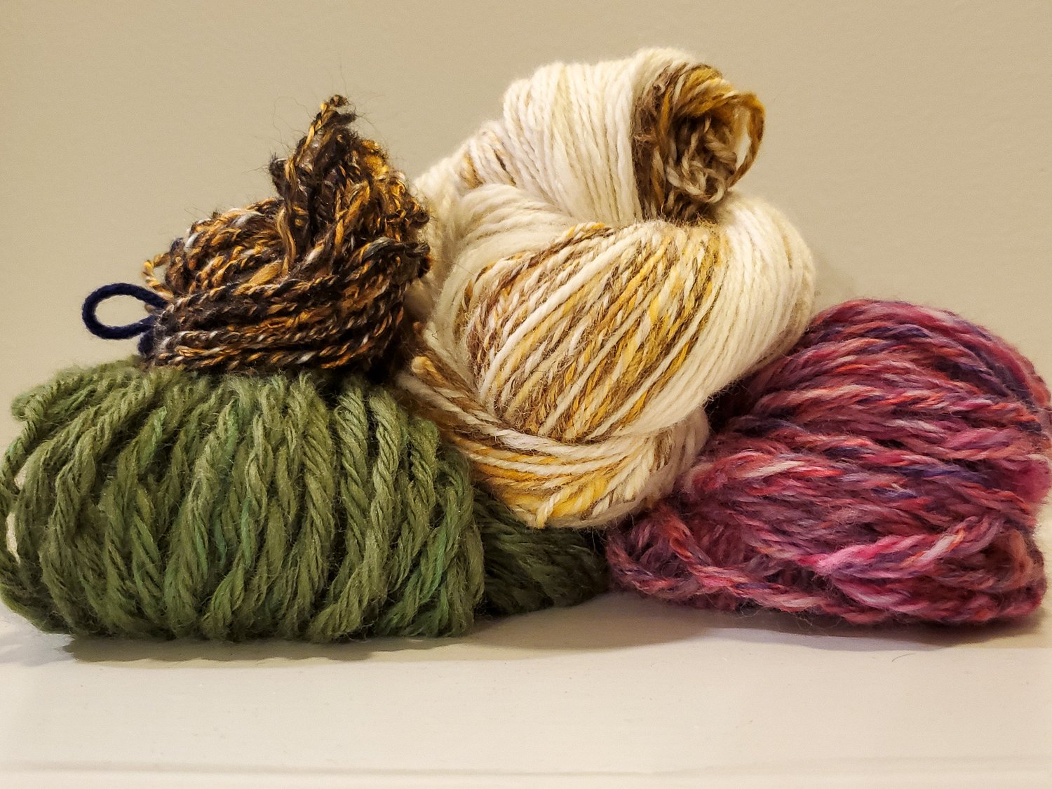 this image shows four handspun skeins of yarn. A pastel fluffy wool, a rich green wool, a thin orange and black, and a cream and brown skein.