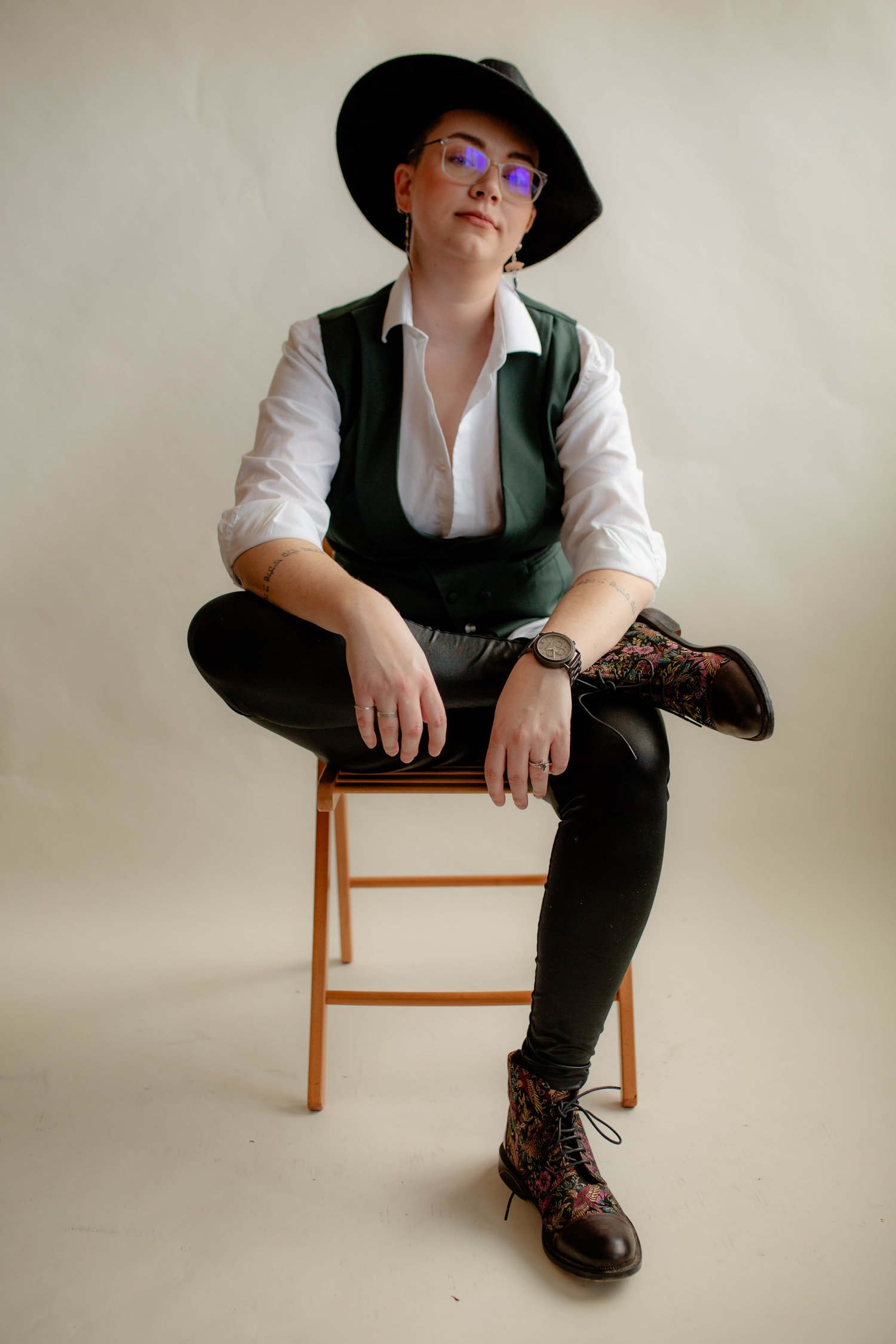 a nonbinary person sits on a chair with one leg crossed over the other, wearing a wide-brimmed black hat, clear-rimmed glasses, a white-button down, a green vest, leather leggings, and embroidered combat boots