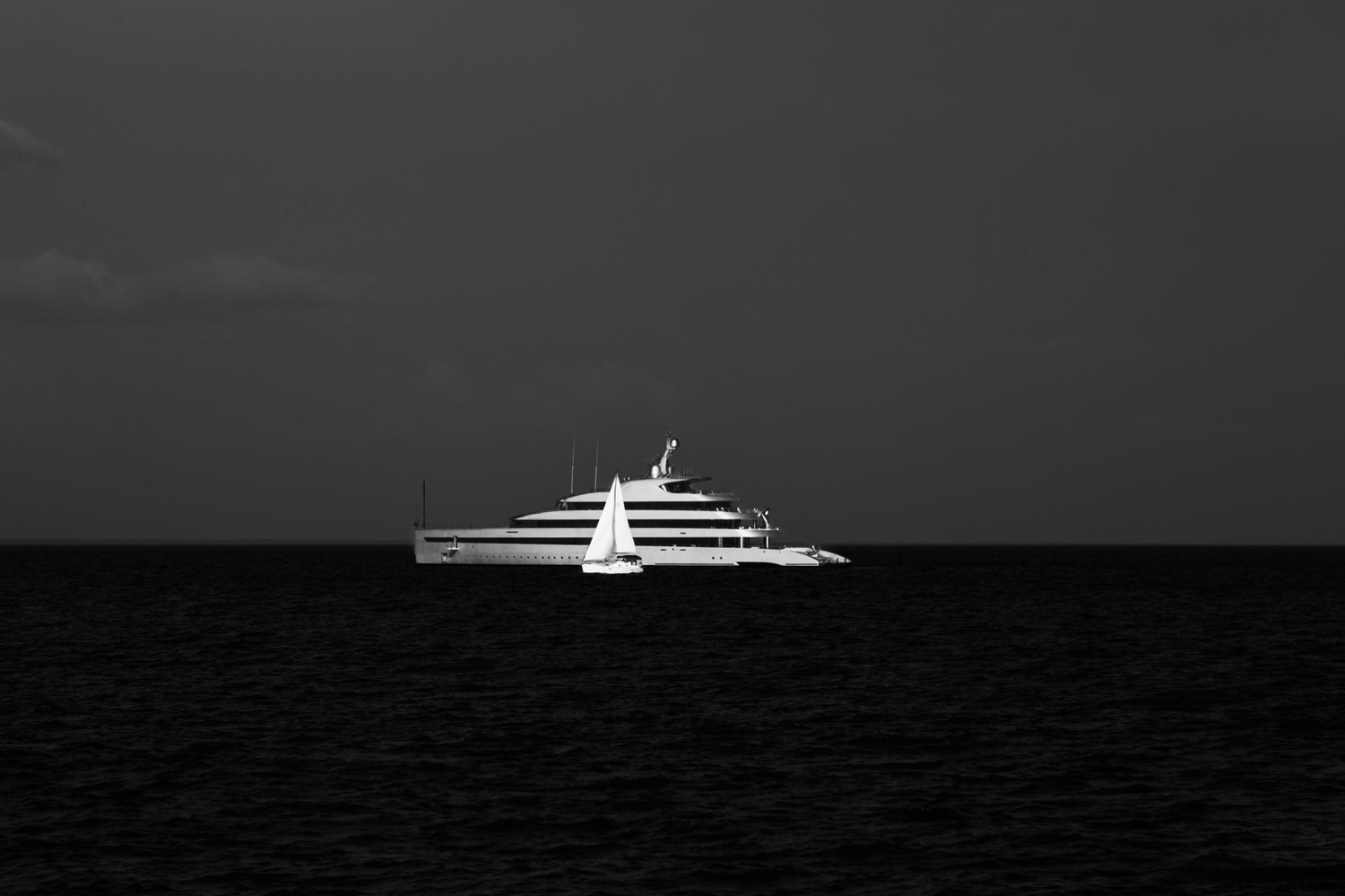 A dramatic black and white horizontal fine art photograph titled Parallel Journeys, featuring a large luxury megayacht and a small white sailboat on a dark sea under a dark sky.
