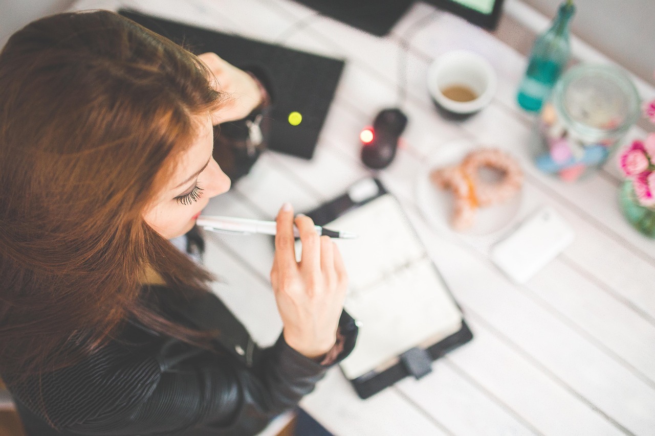 A person sat at a desk sat on the desk is a diary, the person is filling in the diary. there is also a mug and some half eaten food on the desk.