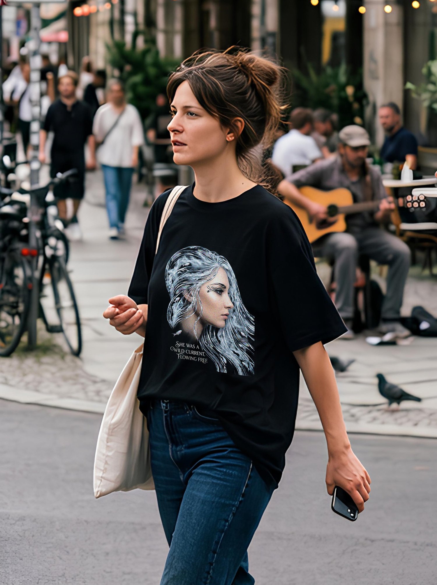 A woman with her hair in a messy bun walks down a city street wearing an oversized black graphic t-shirt and blue jeans. The shirt features a silver-toned illustration of a woman with wave-like flowing hair and the quote "She was a wild current, flowing f