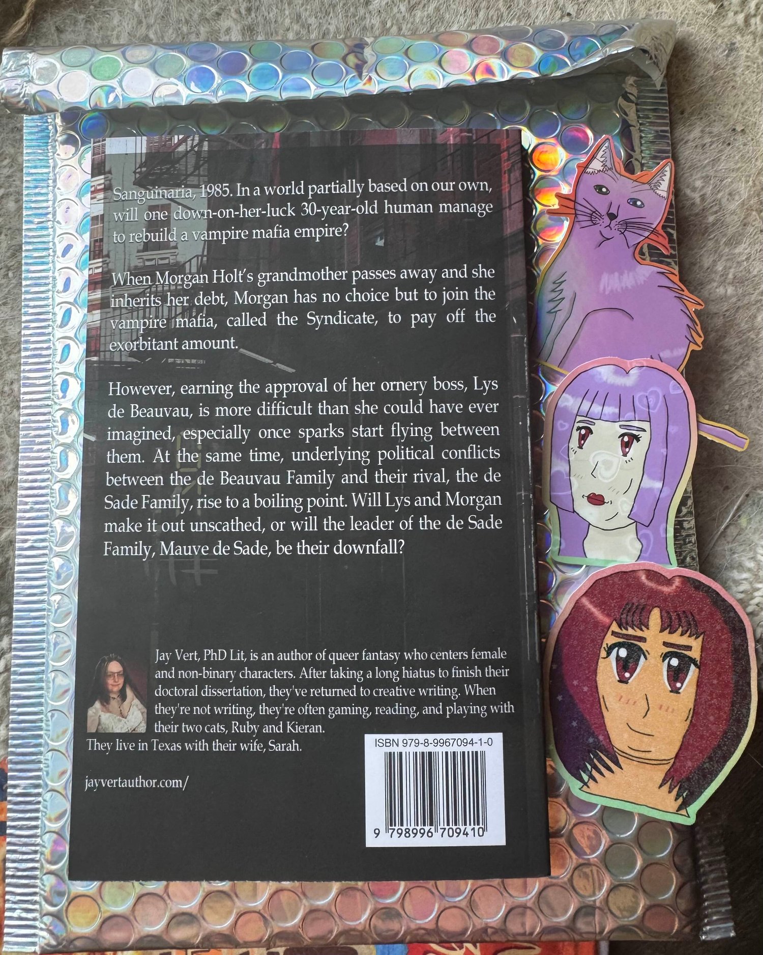 The back cover of a paperback copy of my novella, Blood Sisters, on top of a holographic bubble mailer along with three stickers