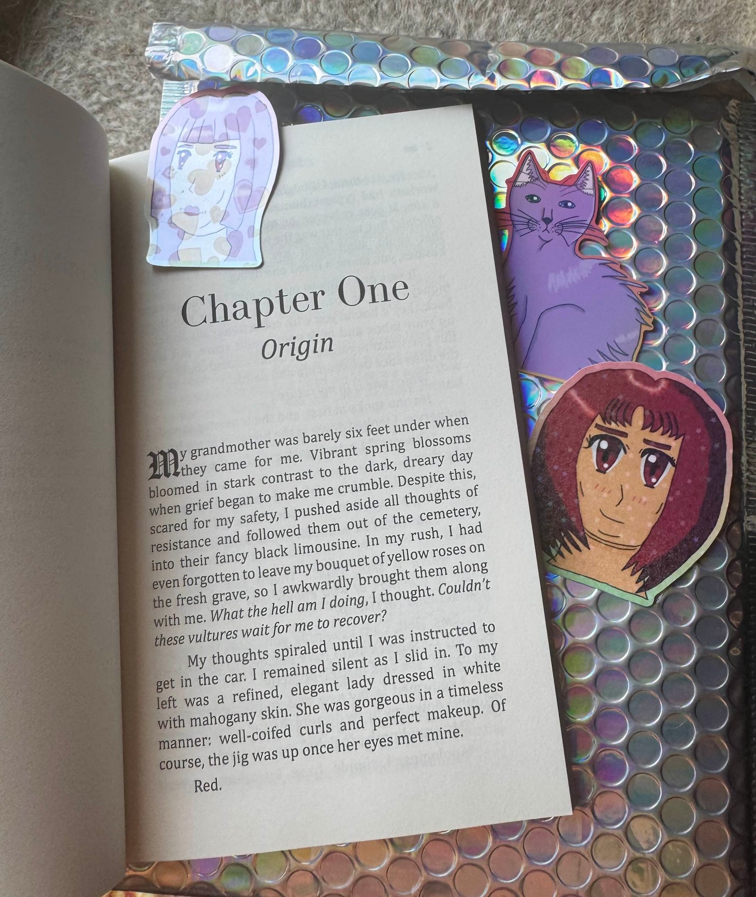 A paperback copy of my novella turned to the first page, Blood Sisters, on top of a holographic bubble mailer along with three stickers