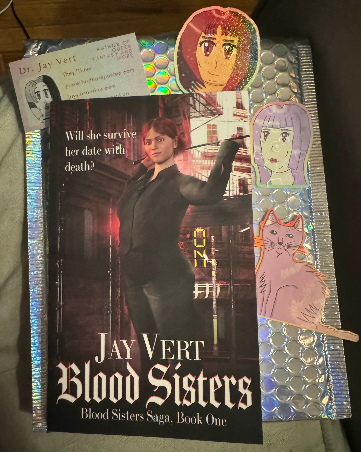 A paperback copy of my novella, Blood Sisters, on top of a holographic bubble mailer along with three stickers