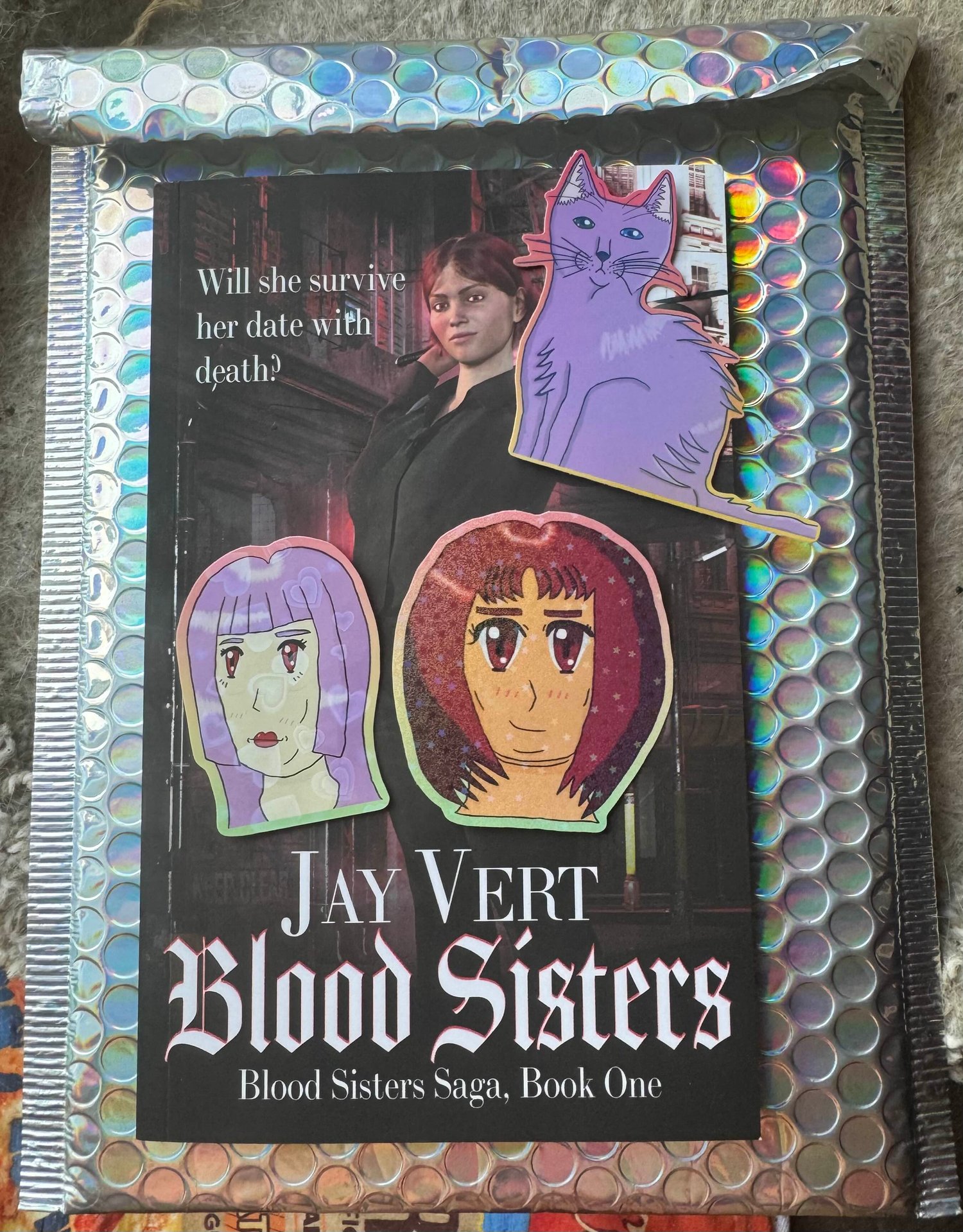 The front cover of a paperback copy of my novella, Blood Sisters, on top of a holographic bubble mailer along with three stickers
