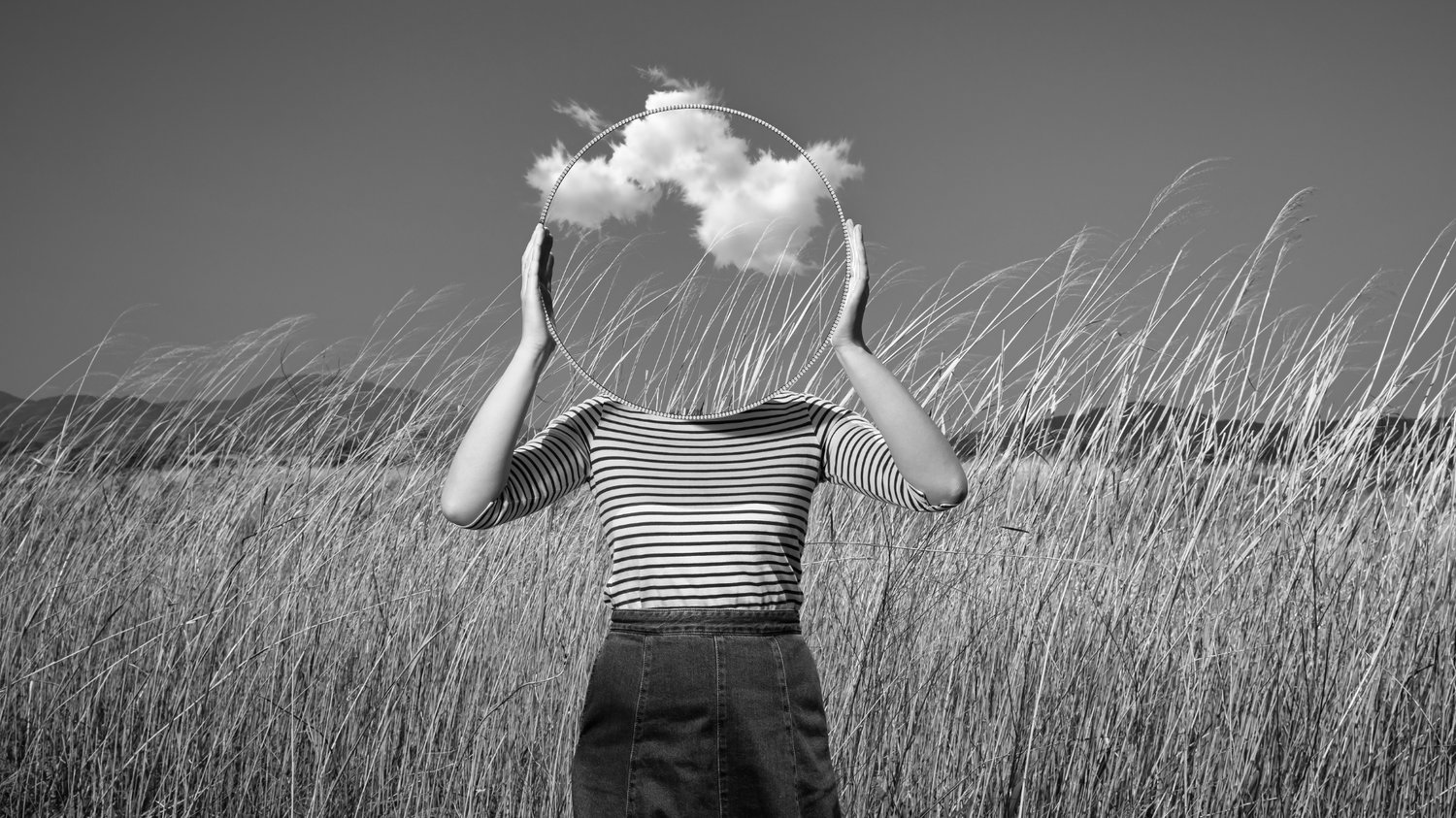 Black-and-white surreal photo of a person standing in a windswept field of tall grass, holding a circular frame overhead where their face would be, with clouds drifting inside the circle — symbolizing the mind caught between old structures and an emerging, open way of thinking.