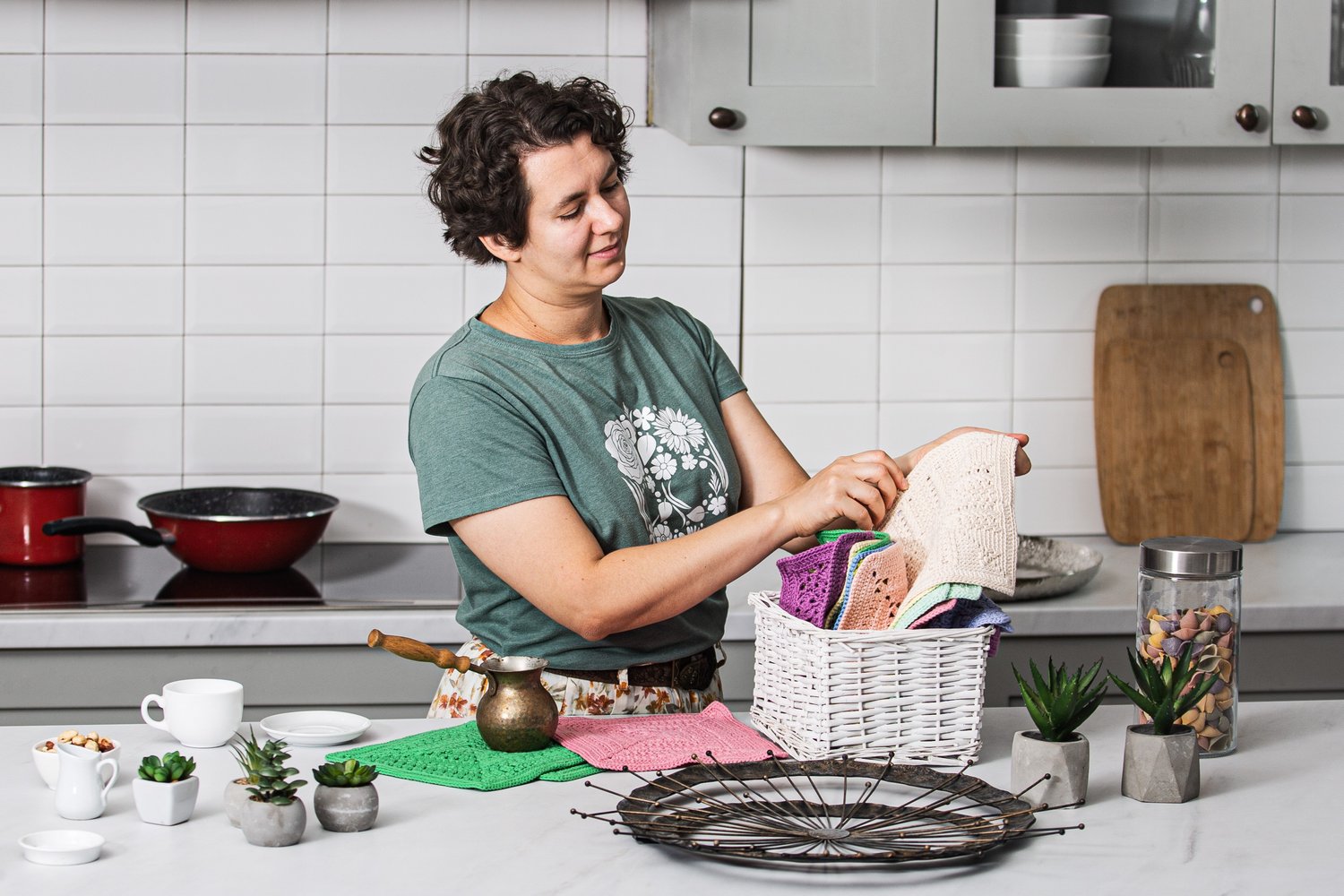 Woman holding a textured beige crochet cotton square from a woven basket of crochet dishcloths in a kitchen. A green DK-weight crochet square with a cezve and dishes sits on the countertop, with a lilac square nearby.