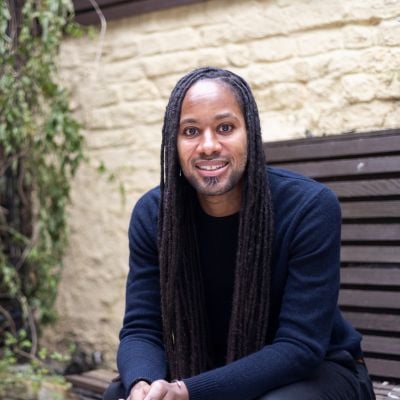 Portrait of Jason Arday seated outdoors on a wooden bench, wearing a dark jumper and smiling warmly at the camera, with a light stone wall and greenery in the background.