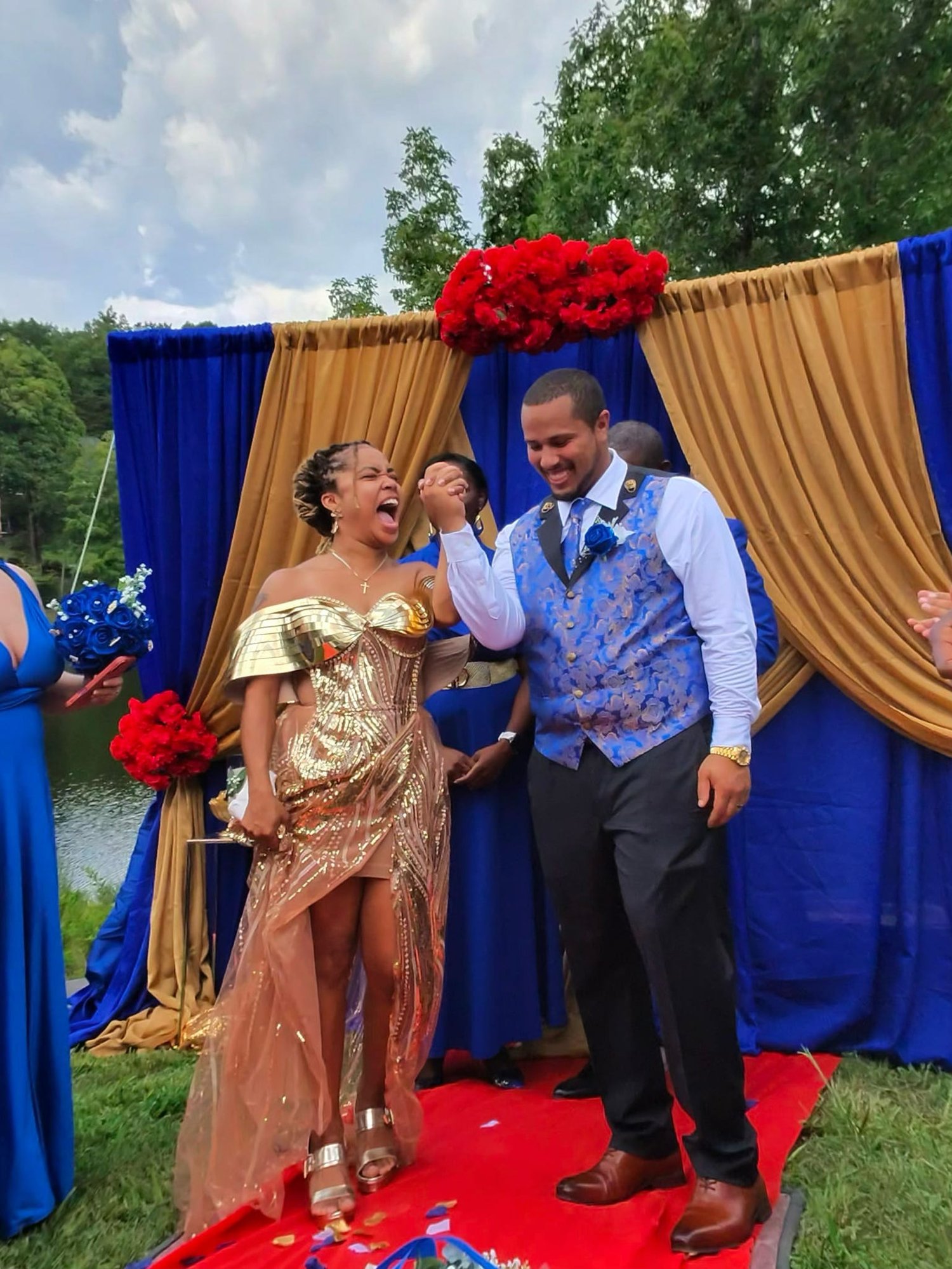 Joyful wedding couple celebrating outdoors with royal blue and gold wedding décor, red florals, and a vibrant personalized ceremony backdrop.