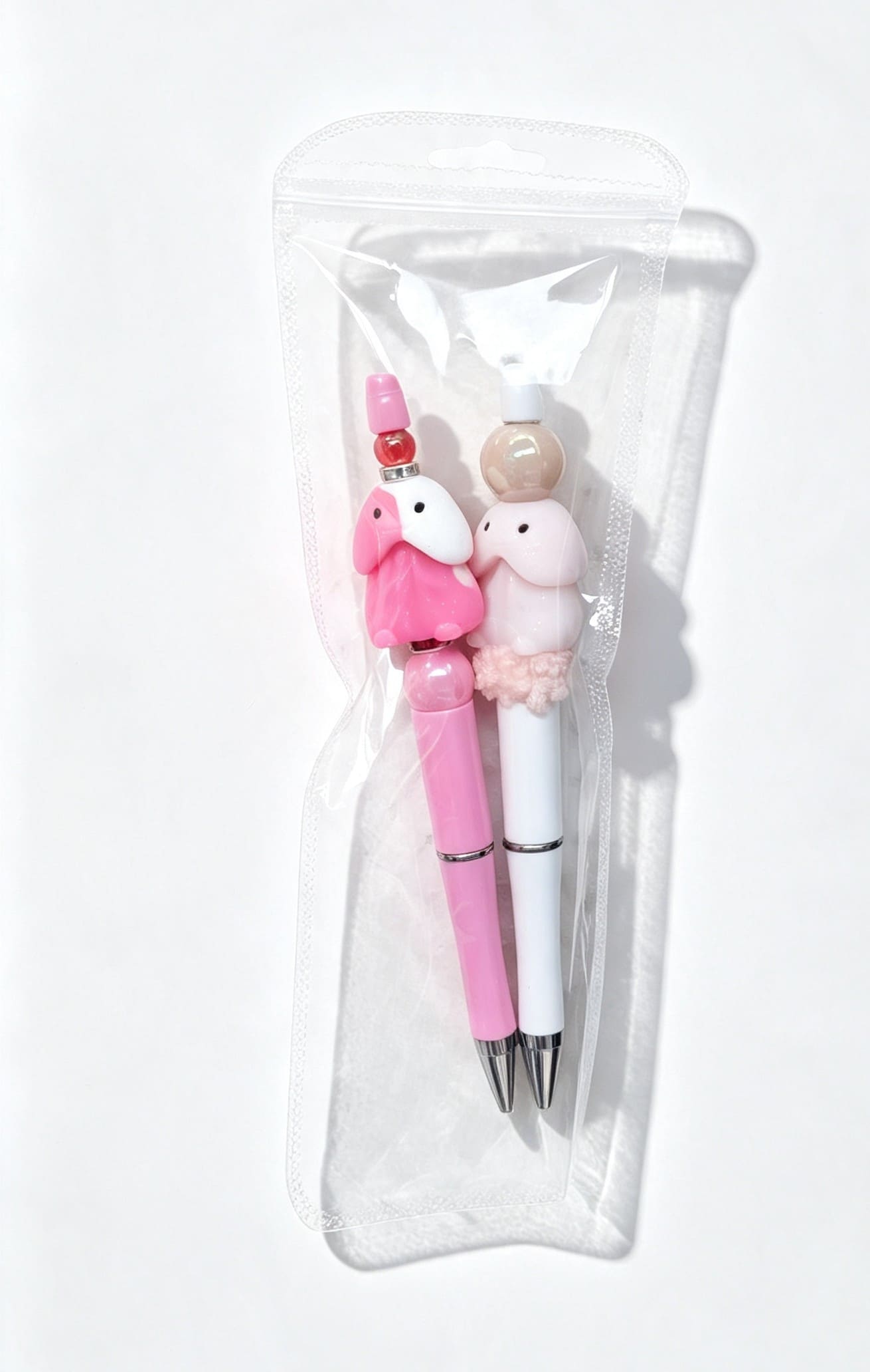 A two-pack set of handmade naughty novelty pens in a clear zipper display pouch, showcasing a pink pen with red rhinestone spacers and a white pen with a custom plush pom-pom texture.