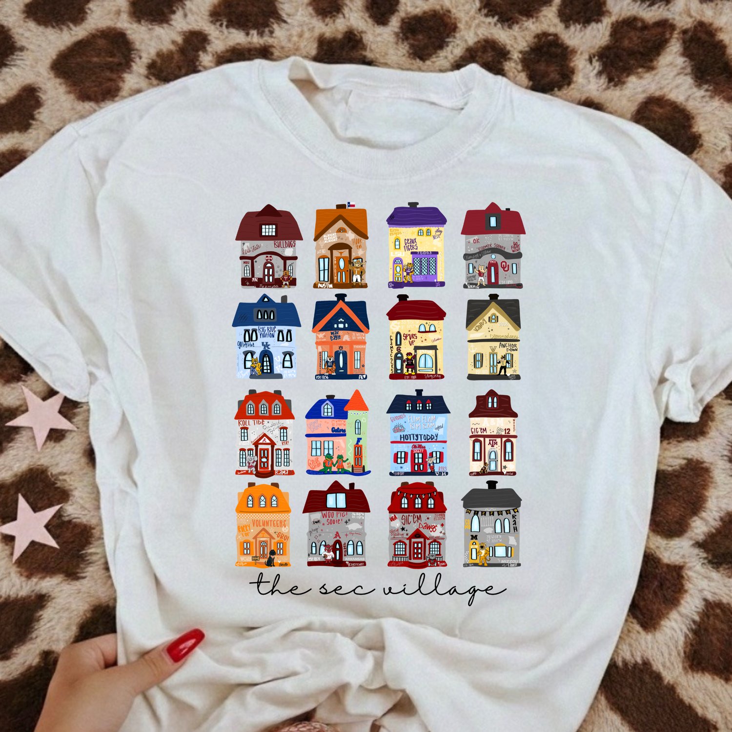 SEC Village T-shirt featuring colorful illustrated college football houses representing SEC teams, printed on a white unisex tee and available as a T-shirt or sweatshirt.