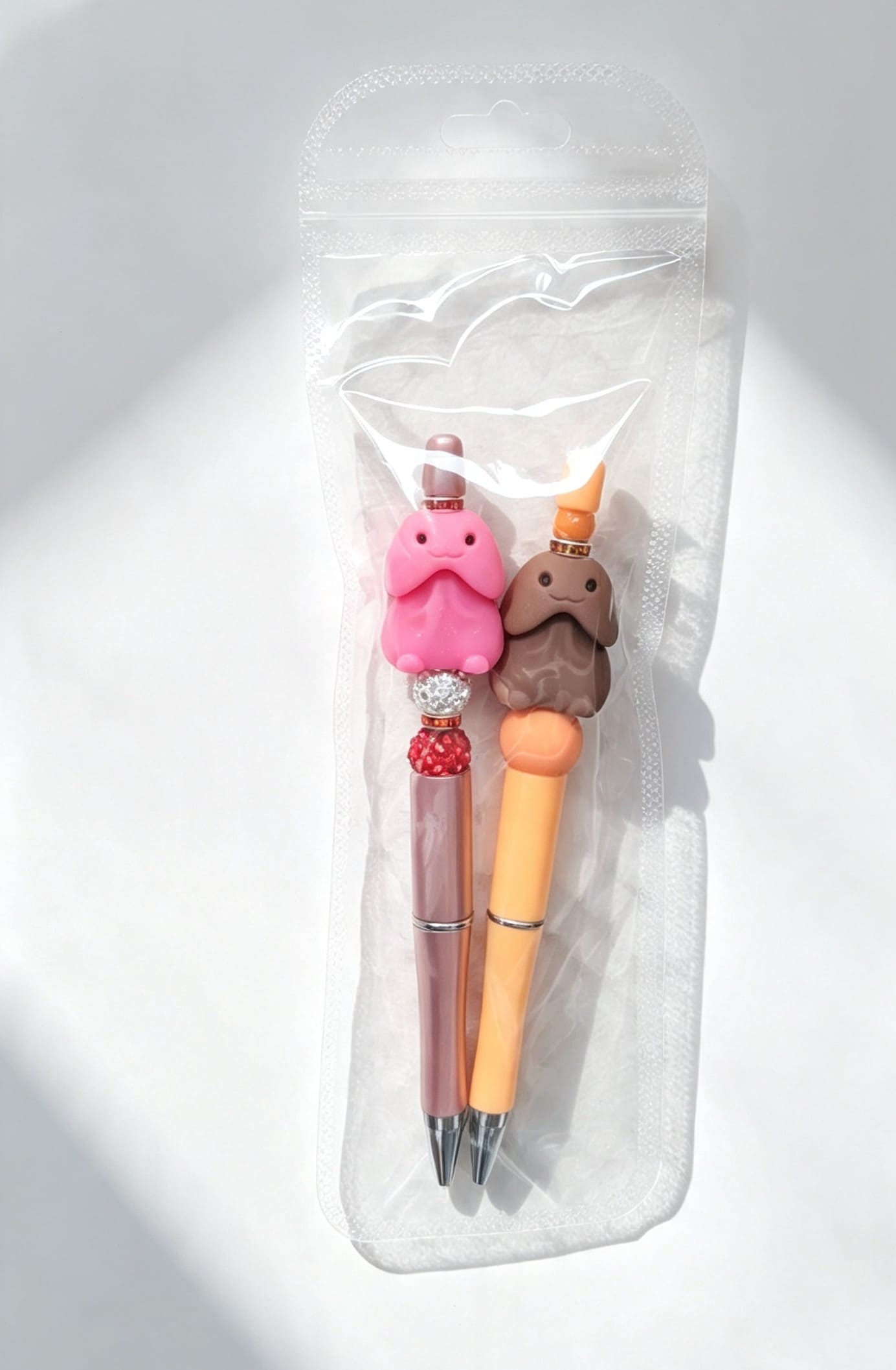 A packaged pair of adult humor beaded pens featuring a metallic rose gold barrel with red glitter balls and an orange barrel with a brown silicone focal bead, sealed in clear protective packaging.