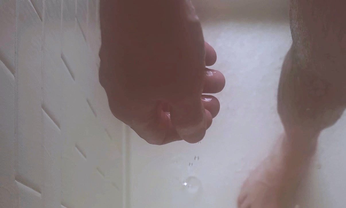 Close-up of a PRiCE’s hand and leg in a shower, partially obscured by water, photographed from a low angle against white tile.