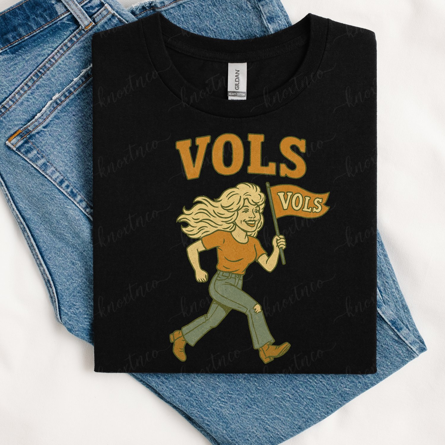 Black Tennessee Volunteers T-shirt featuring a vintage Dolly-inspired blonde woman carrying an orange Vols flag with retro VOLs lettering, styled with blue jeans.