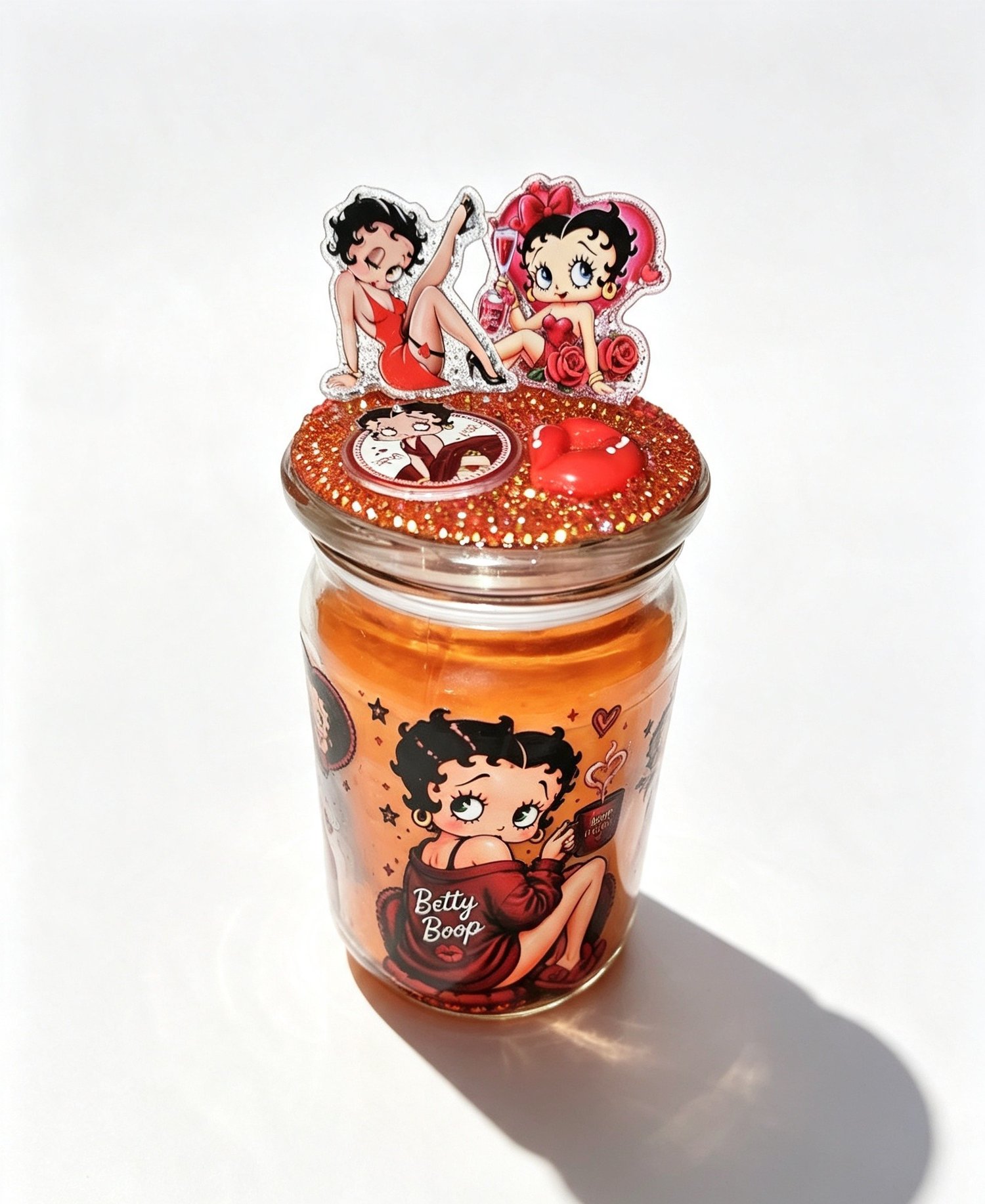 Top view of a handmade Betty Boop candle lid covered in orange glitter and rhinestones with 3D acrylic character cutouts and a red lip charm.