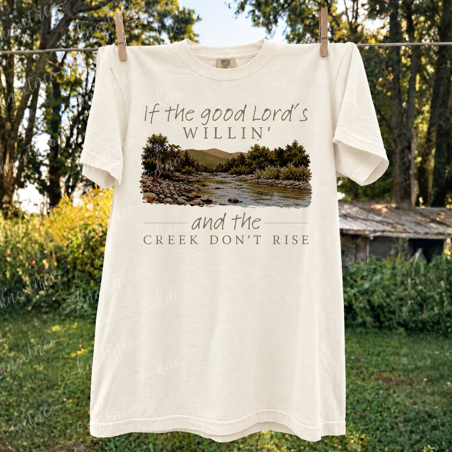 If the Good Lord’s willin’ and the creek don’t rise Christian Southern saying design with a vintage mountain creek scene, shown on a comfort colors ivory tshirt.