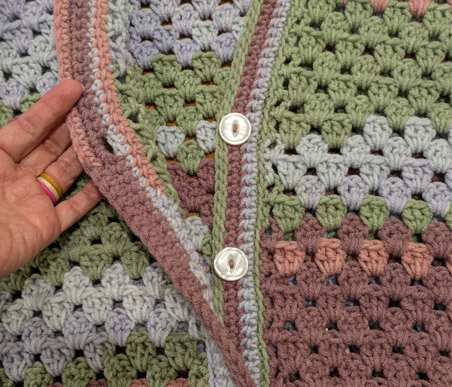 Close-up of a handmade granny stitch crochet cardigan in soft green, dusty rose, and pale blue, showing the textured button band, white buttons, and multicolor granny stitch pattern.
