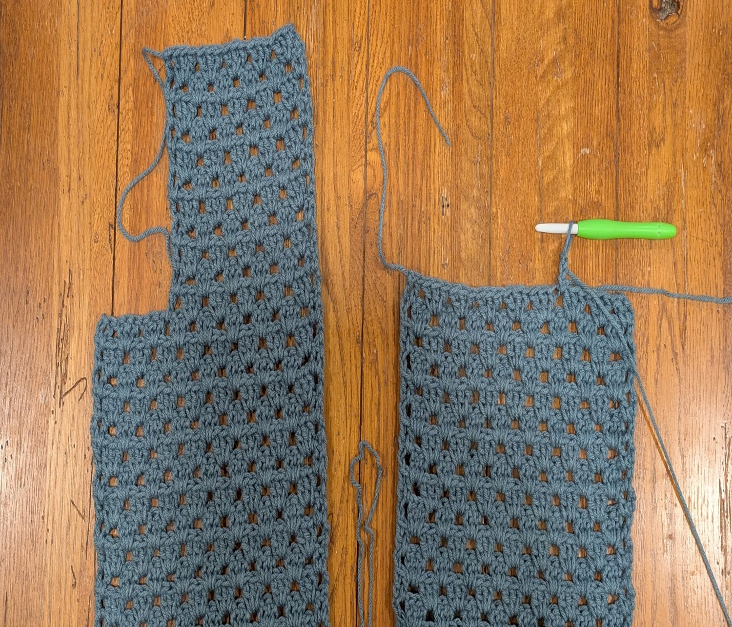 Two granny stitch crochet cardigan panels in progress using blue-gray yarn, shown with a green-handled crochet hook on a wooden surface, demonstrating handmade sweater construction and custom sizing.
