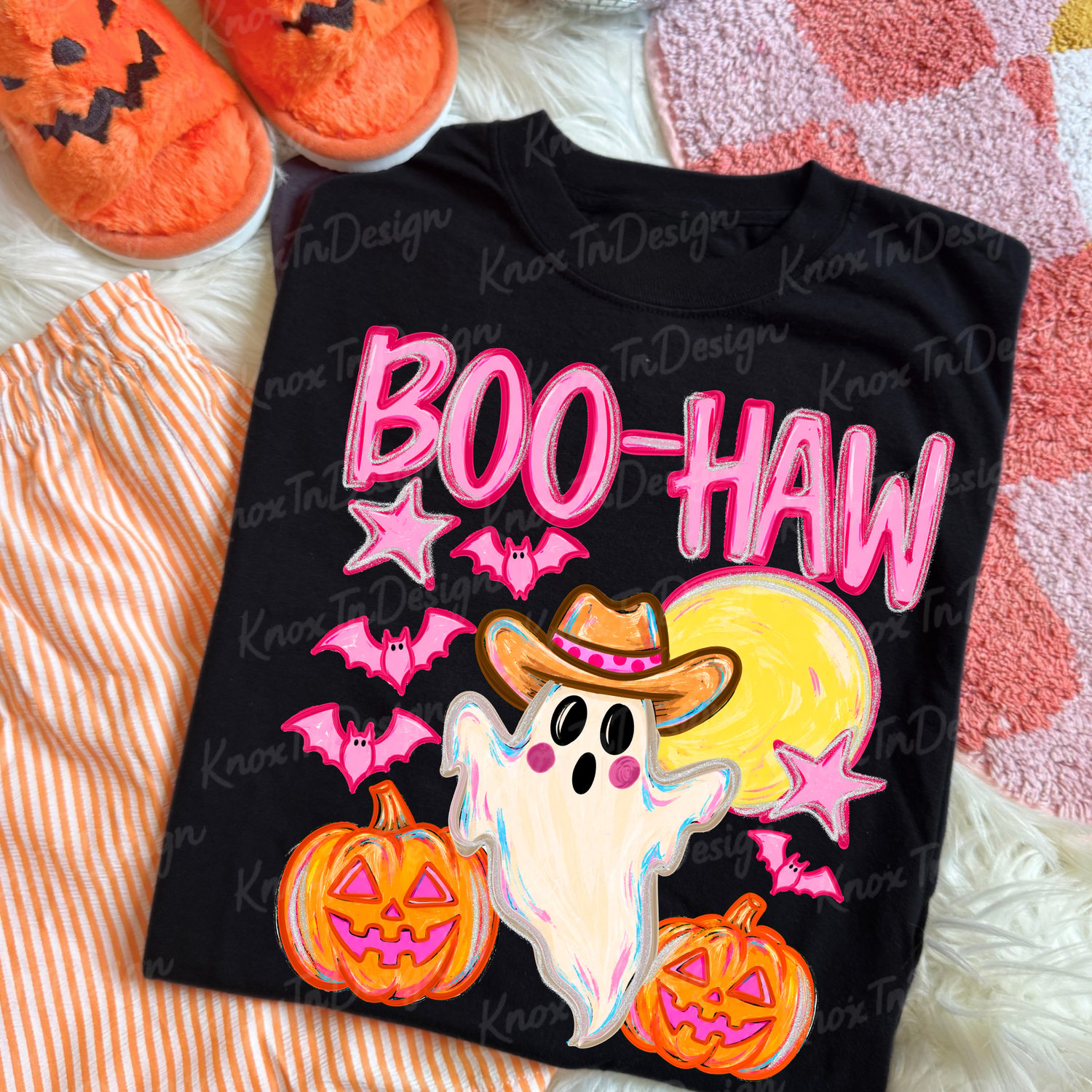 Front view of a white Boo-Haw western Halloween t-shirt featuring a cartoon ghost wearing a cowgirl hat flanked by pink-faced pumpkins, bats, and stars, photographed in an outdoor pumpkin patch.